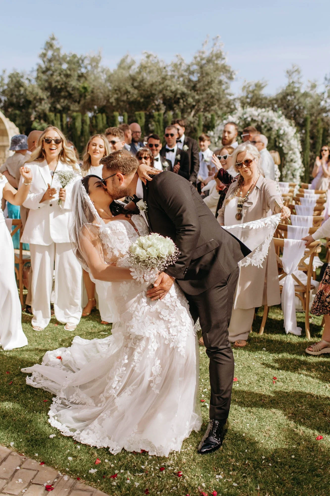A newlywed couple shares a kiss during their outdoor wedding ceremony, surrounded by friends and family on a sunny day with blossoming trees in the background.