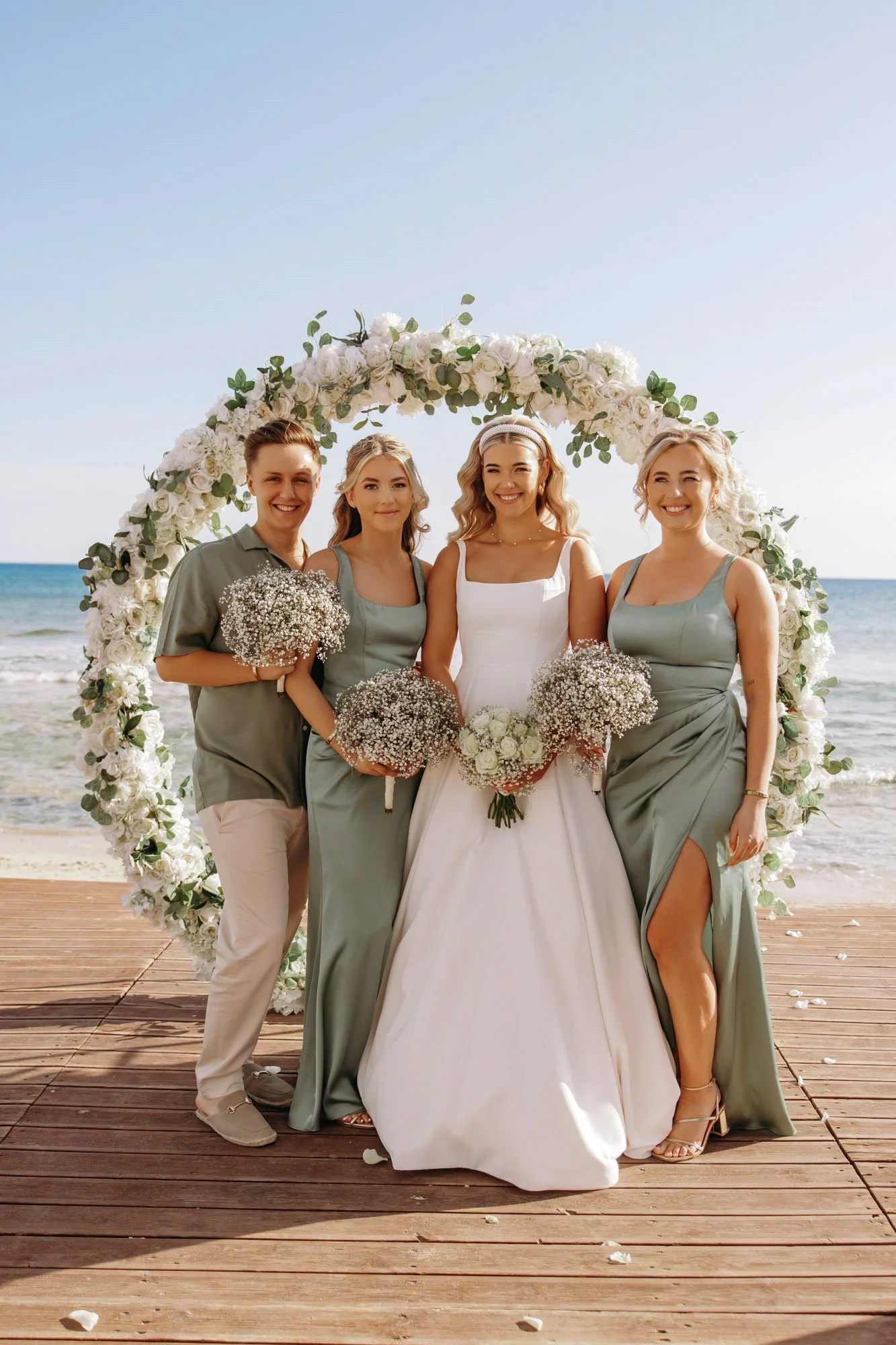 A bride and three bridesmaids standing on a wooden platform by the beach during a wedding, with a large floral arch behind them, all smiling and holding bouquets.