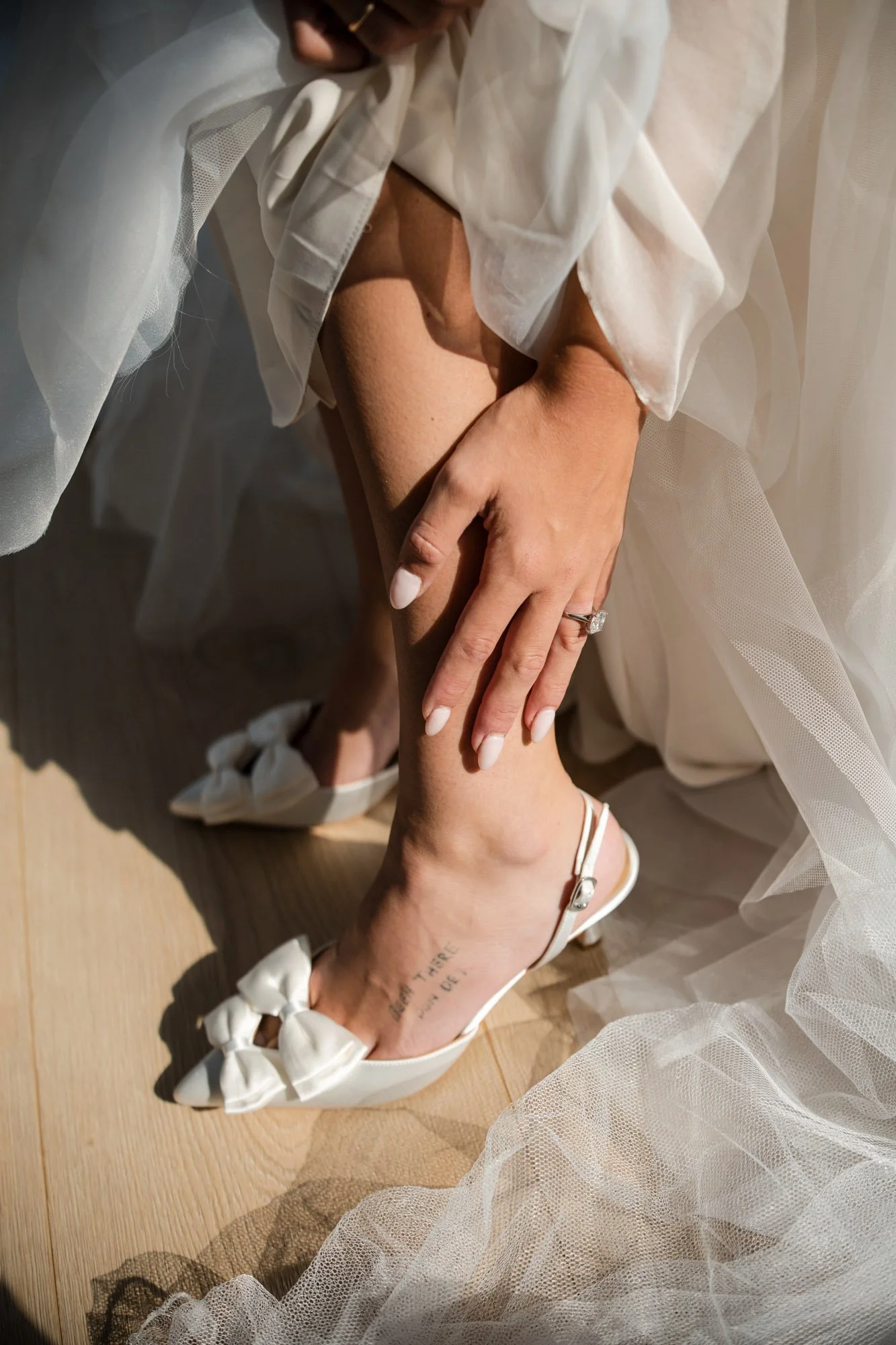 Close-up of a woman wearing white high heels with bows, a white tattoo on her ankle, a clear ring on her finger, a white sheer dress, and she is touching her leg.