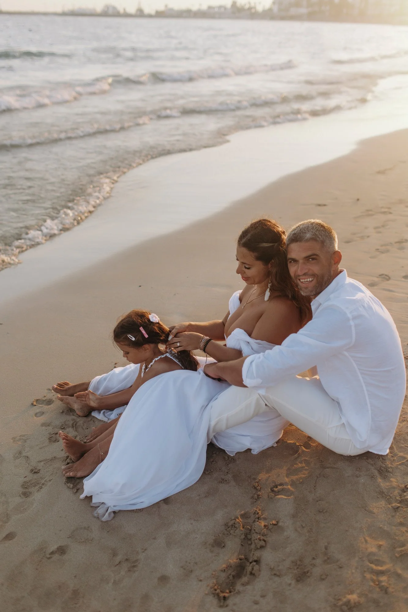 A family sitting on the sandy beach during sunset, dressed in white clothes, enjoying a relaxed moment by the shoreline.