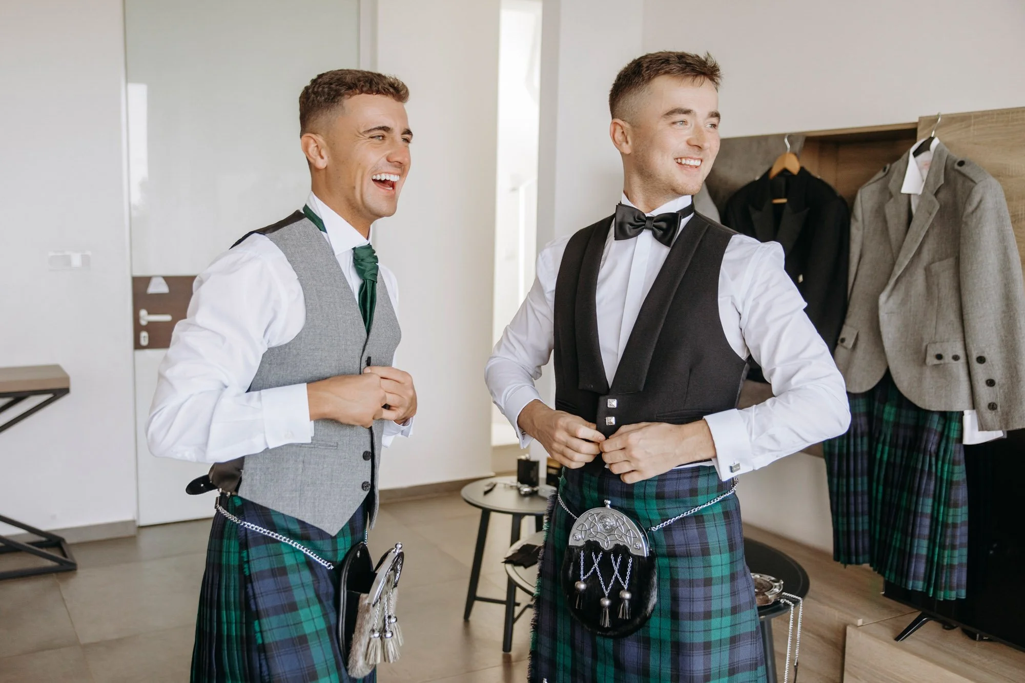 Two young men wearing traditional Scottish kilts and formal attire, adjusting their vests and tuxedos, preparing for a special event in a dressing room.
