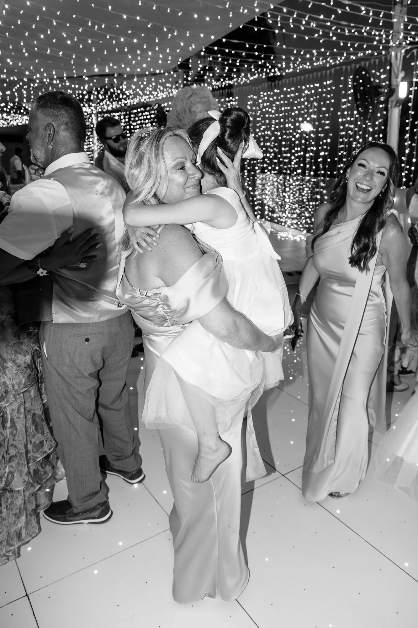 People celebrating at a party or wedding reception with fairy lights overhead, some dressed in formal attire, with joy and laughter.