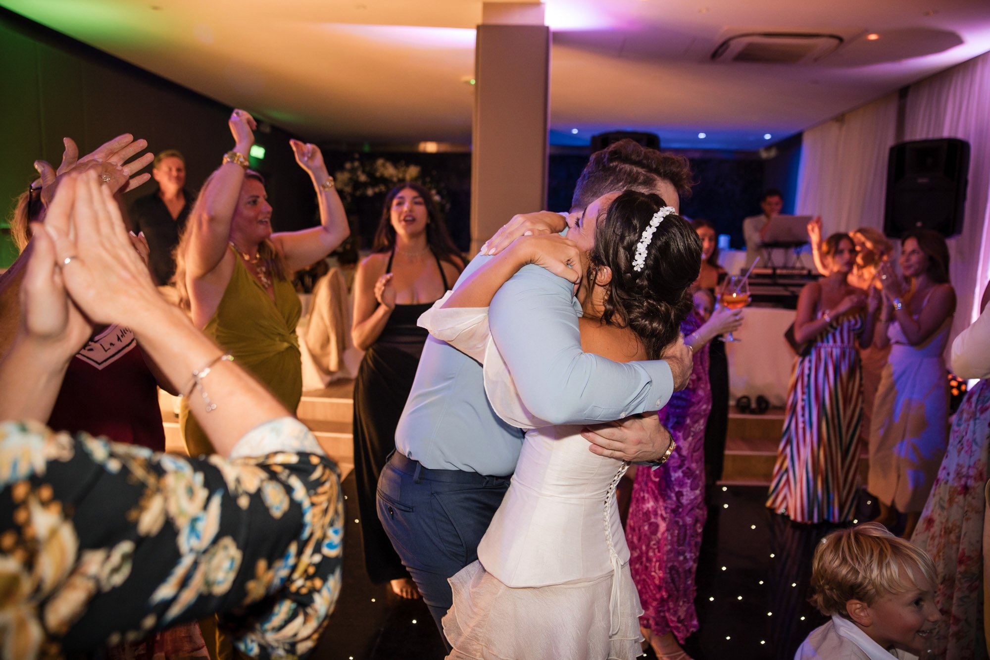 A wedding reception with guests dancing and celebrating as a bride and groom embrace in the center of the dance floor, surrounded by friends and family.