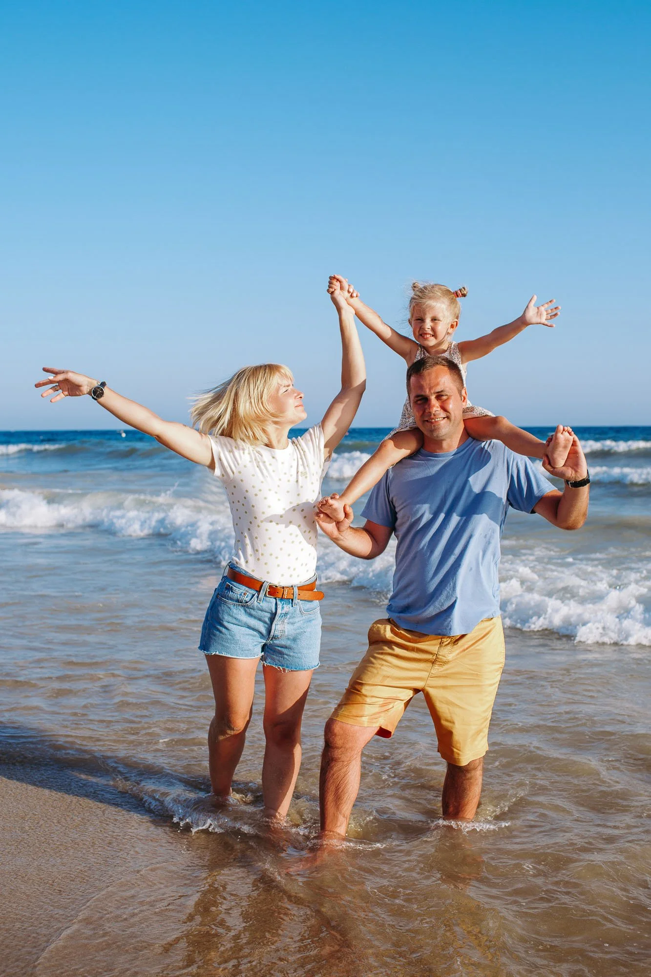 A happy family enjoying time at the beach, with a woman, a man, and a young girl standing in the shallow water, holding hands and smiling.