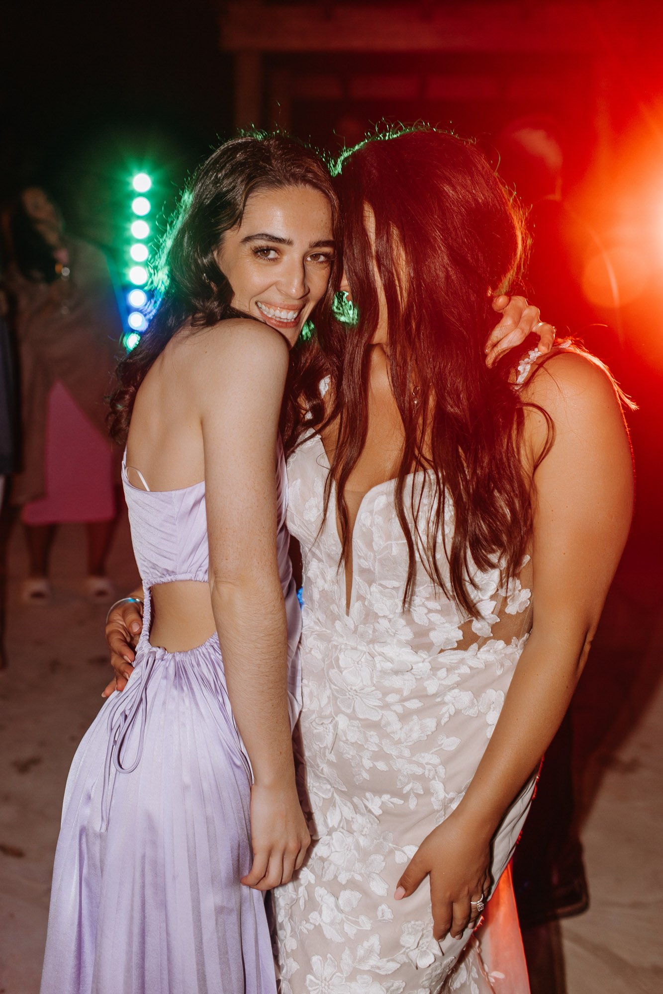 Two women wearing elegant dresses dancing at a party or celebration with colorful lights in the background.
