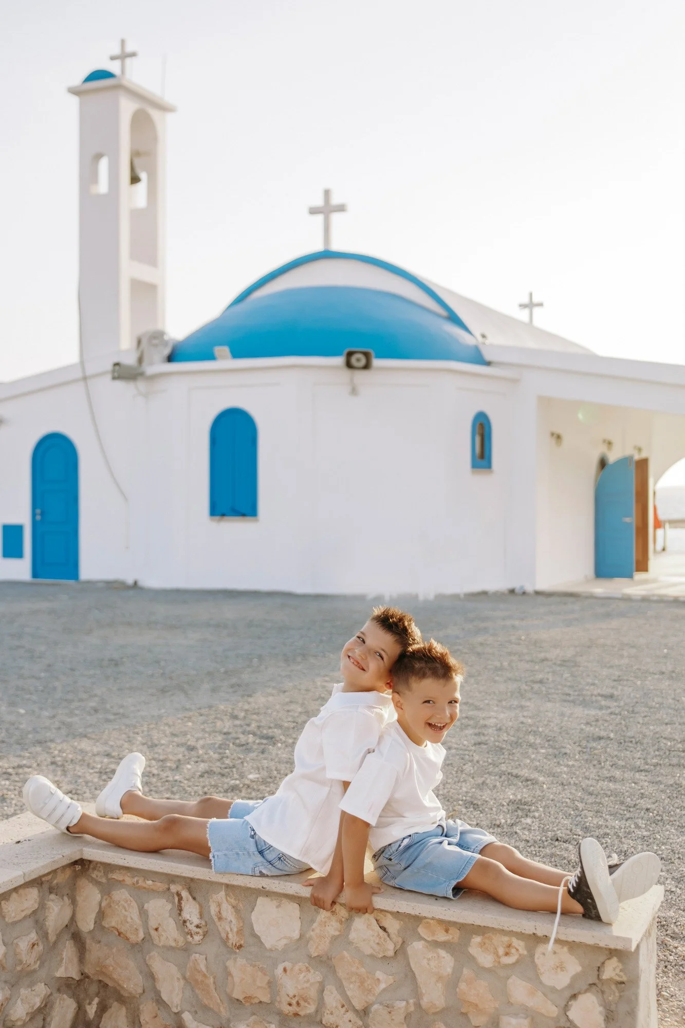 Two smiling boys in white shirts and shorts sit on a stone wall in front of a white and blue Greek Orthodox church with crosses on its roof, during daytime.