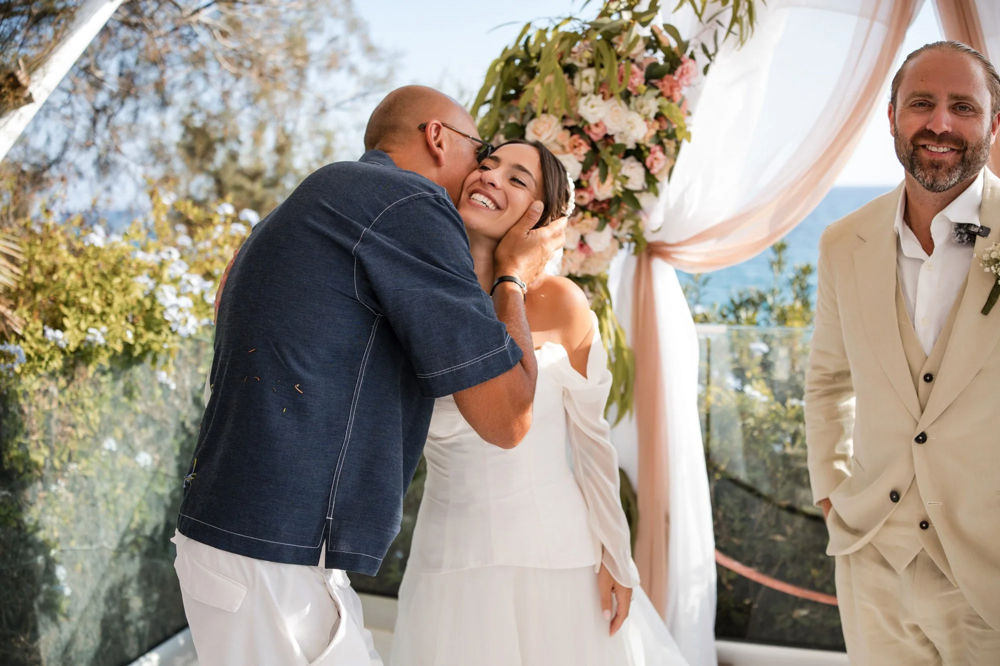 A groom in a beige suit smiling at the wedding ceremony, a bride in a white off-shoulder dress smiling as an officiant kisses her on the cheek, with a floral backdrop and ocean views in the background.