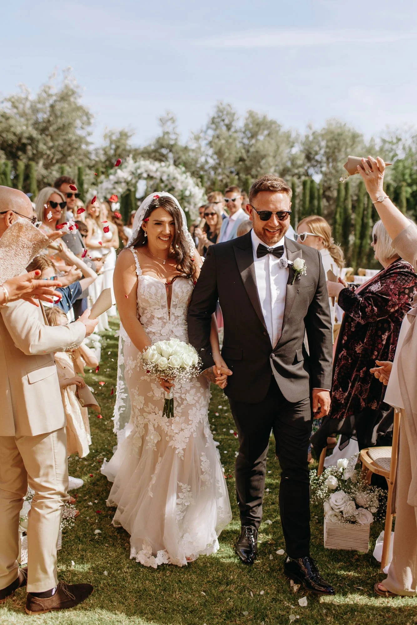 A bride and groom walking outdoors after their wedding ceremony, surrounded by friends and family throwing flower petals, on a sunny day.