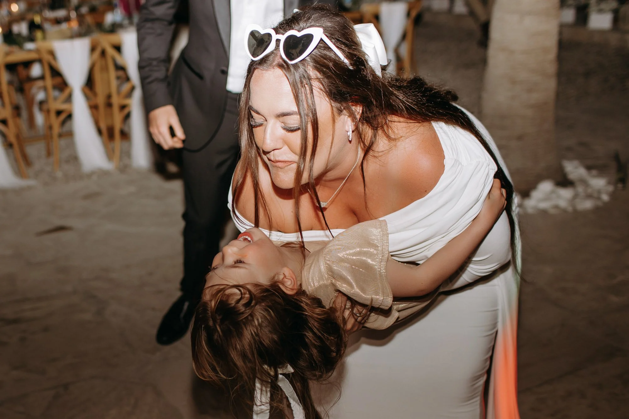 A woman in a white dress is holding a young girl in a gold dress as they dance together at an outdoor celebration or wedding, with wooden chairs and a person in a suit in the background.