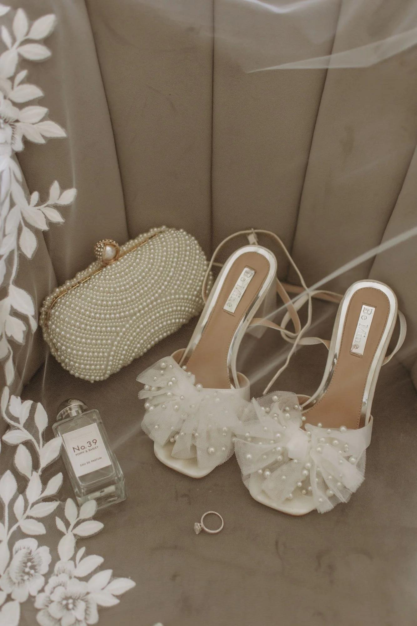 A pair of white high-heeled shoes with pearl and lace decorations, a pearl-embellished clutch purse, a bottle of perfume, and a ring display on a cushioned surface, likely for a wedding or special event.