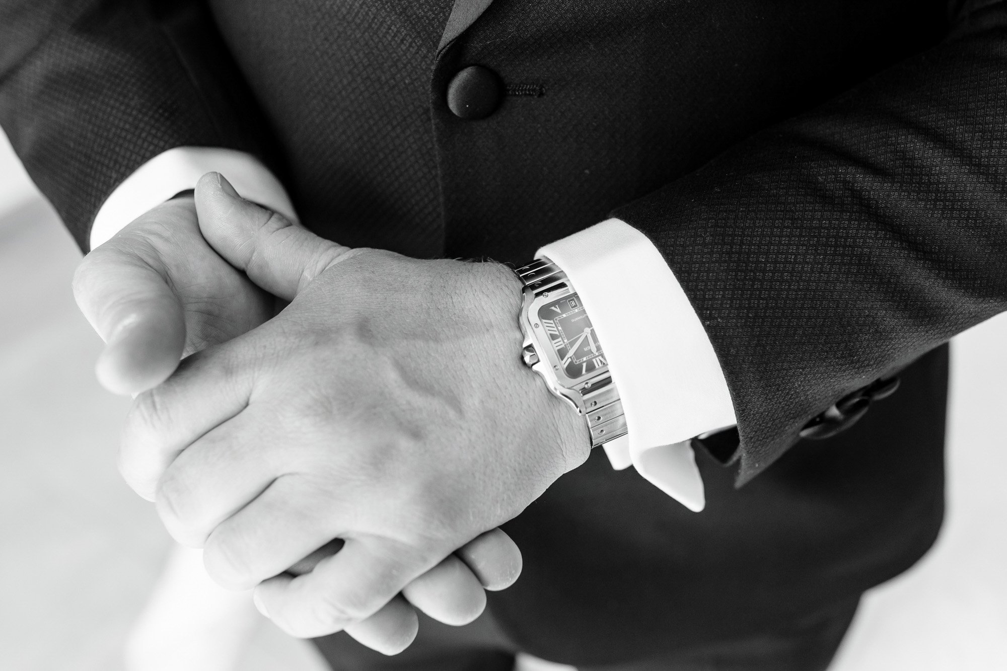 Close-up of a man's hands clasped, wearing a dark suit with white shirt cuffs and a wristwatch.