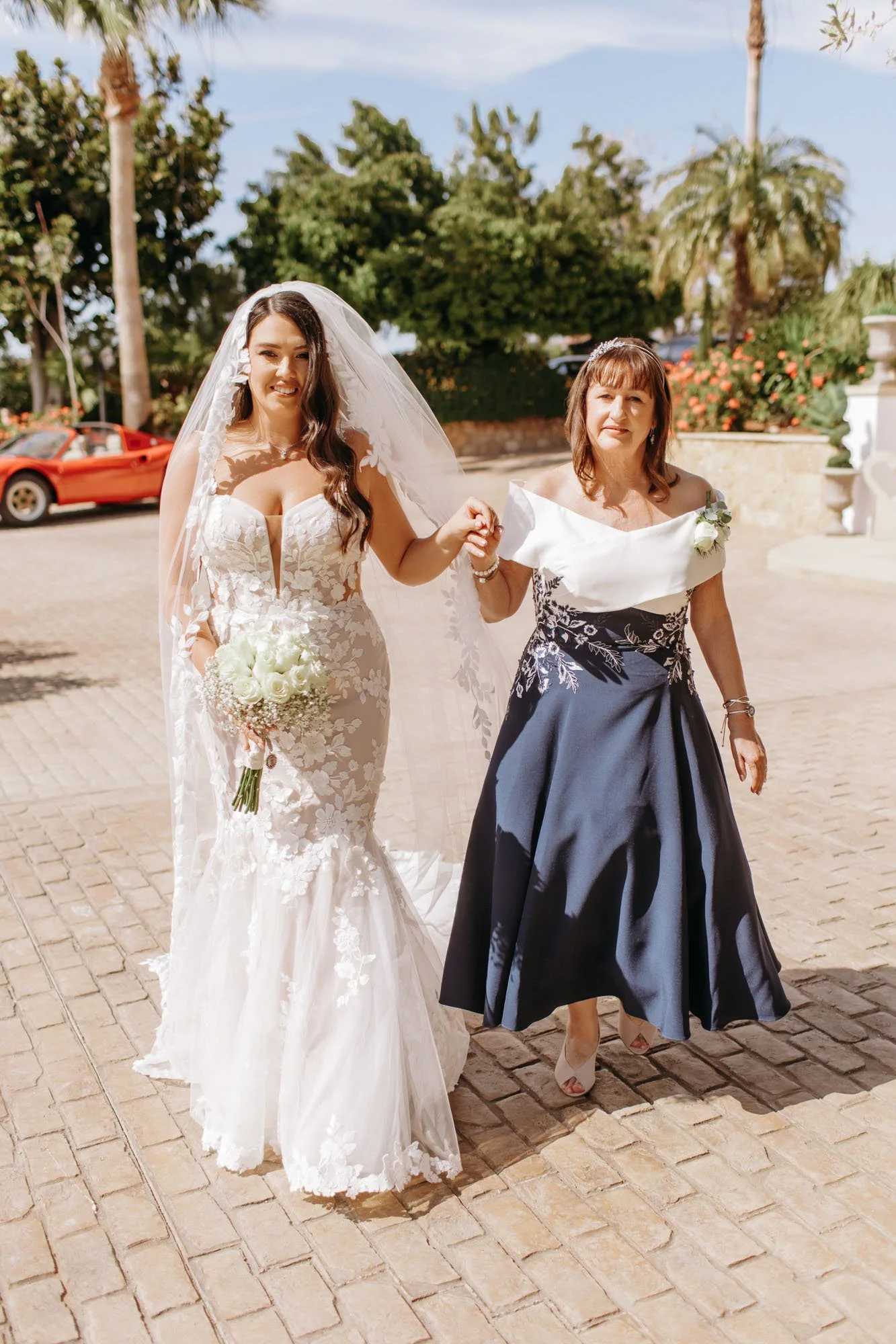 A bride in a white lace wedding dress with a veil holding a bouquet walking with a woman in a navy skirt and white top outside on a sunny day.