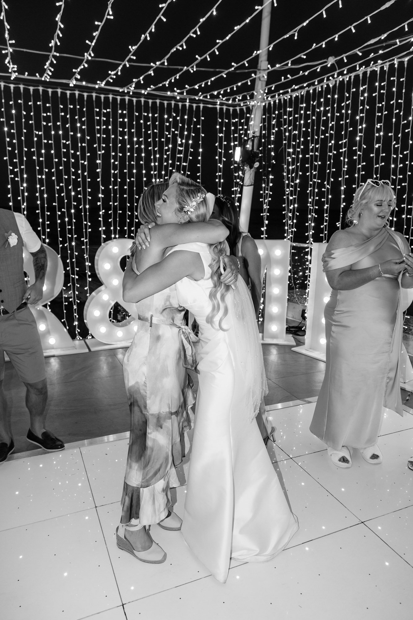 Two women hugging at a celebration event with fairy lights and illuminated letters in the background.