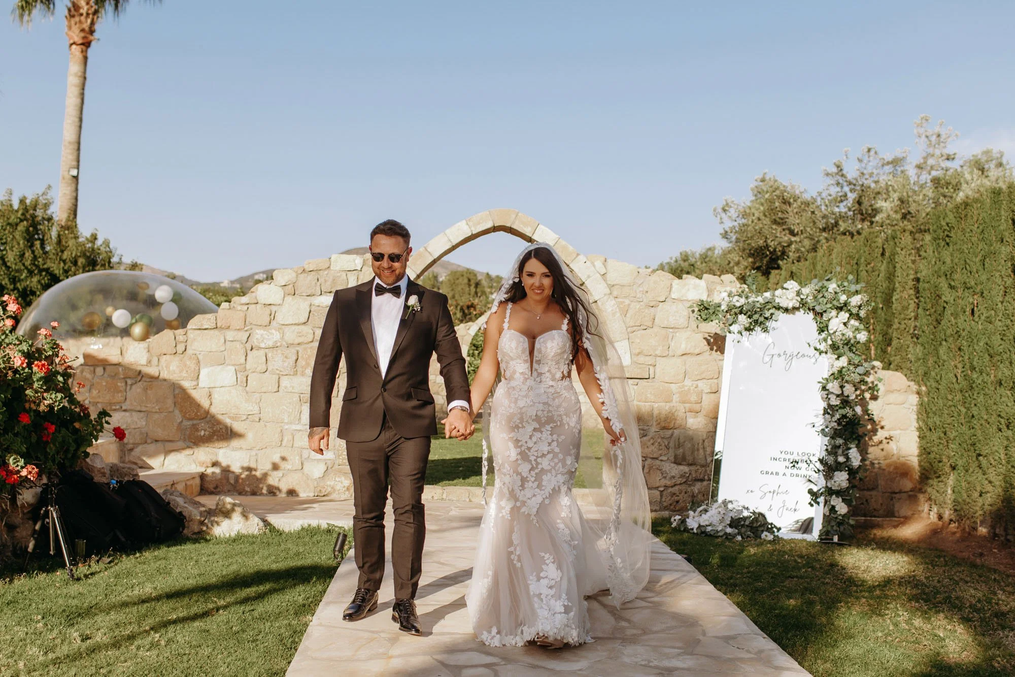 A bride and groom walking hand in hand outdoors, smiling, with a stone arch and decorated sign in the background at a wedding ceremony.
