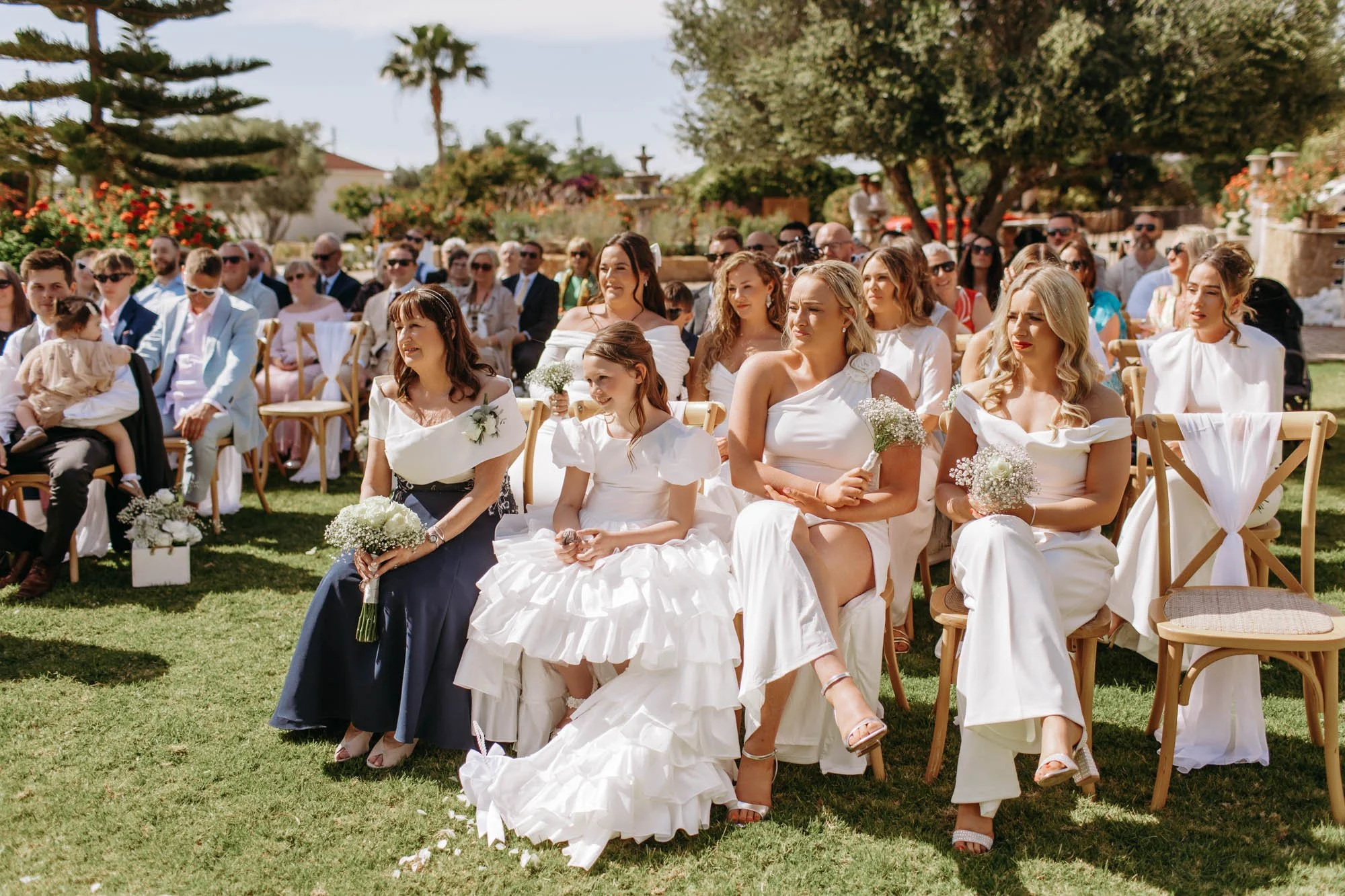 Wedding guests seated outdoors during the ceremony, dressed in formal attire, with many women holding bouquets and kids present.