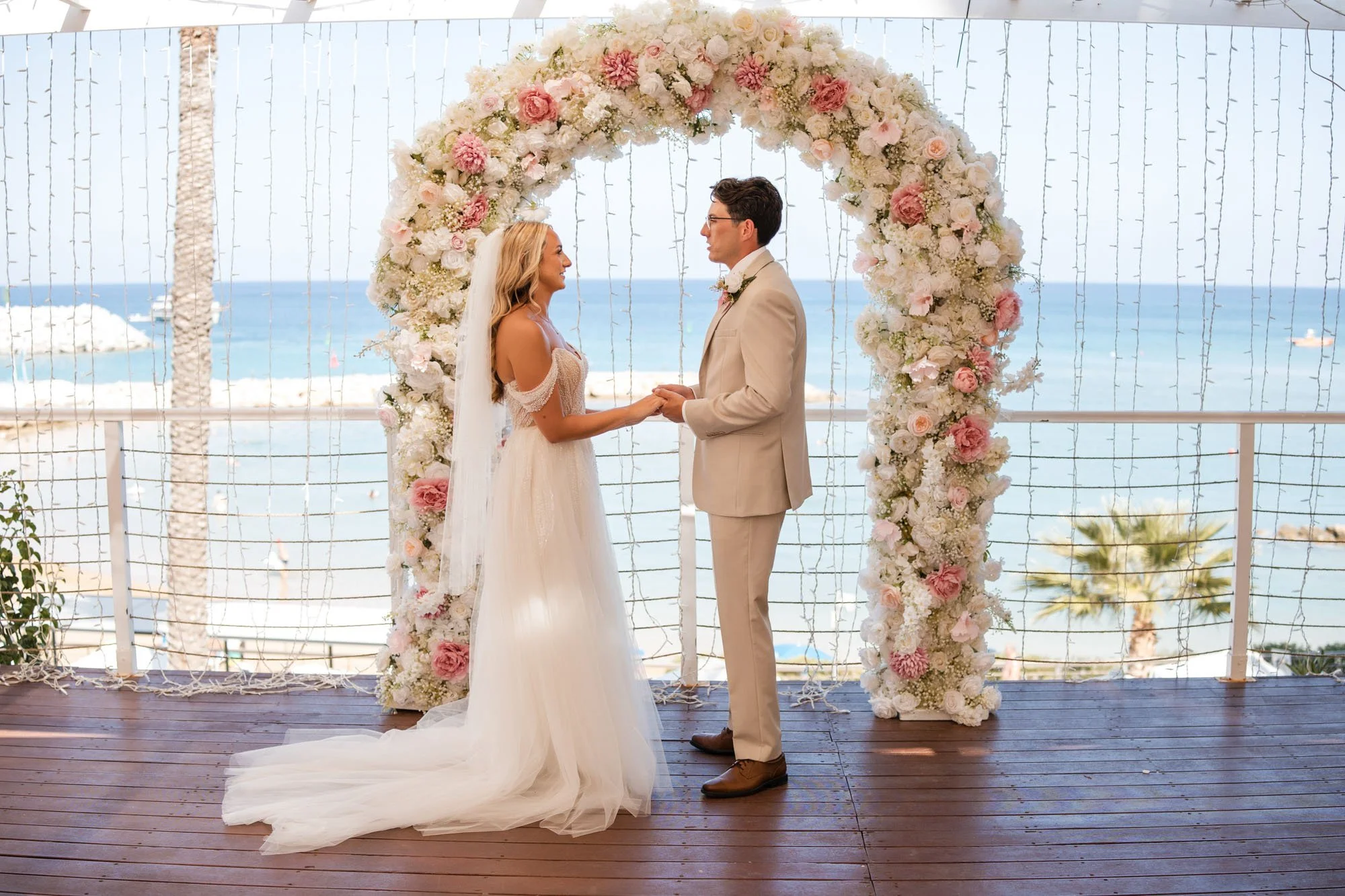 A bride and groom holding hands during their wedding ceremony on a beachside balcony, with a floral arch and ocean view in the background.