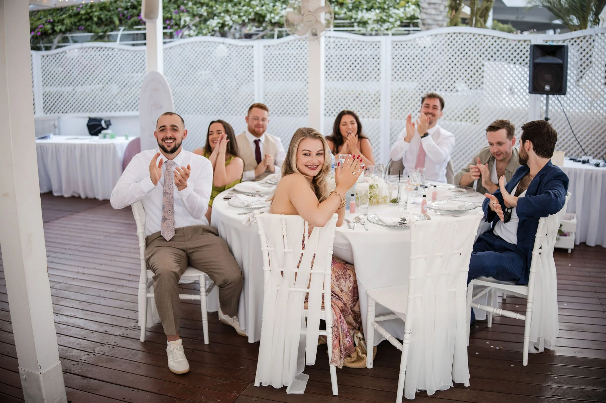 A group of people at a wedding reception sitting around a table, smiling and clapping, with a white lattice fence and greenery in the background.