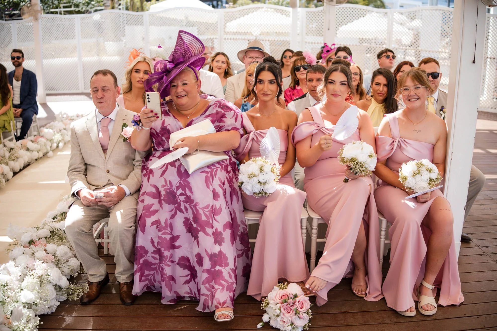 A group of wedding guests seated on chairs, including women in pink dresses holding bouquets, a lady in a pink and purple patterned dress with a large purple hat taking a photo, and men in suits, attending a wedding ceremony on a decorated outdoor ve