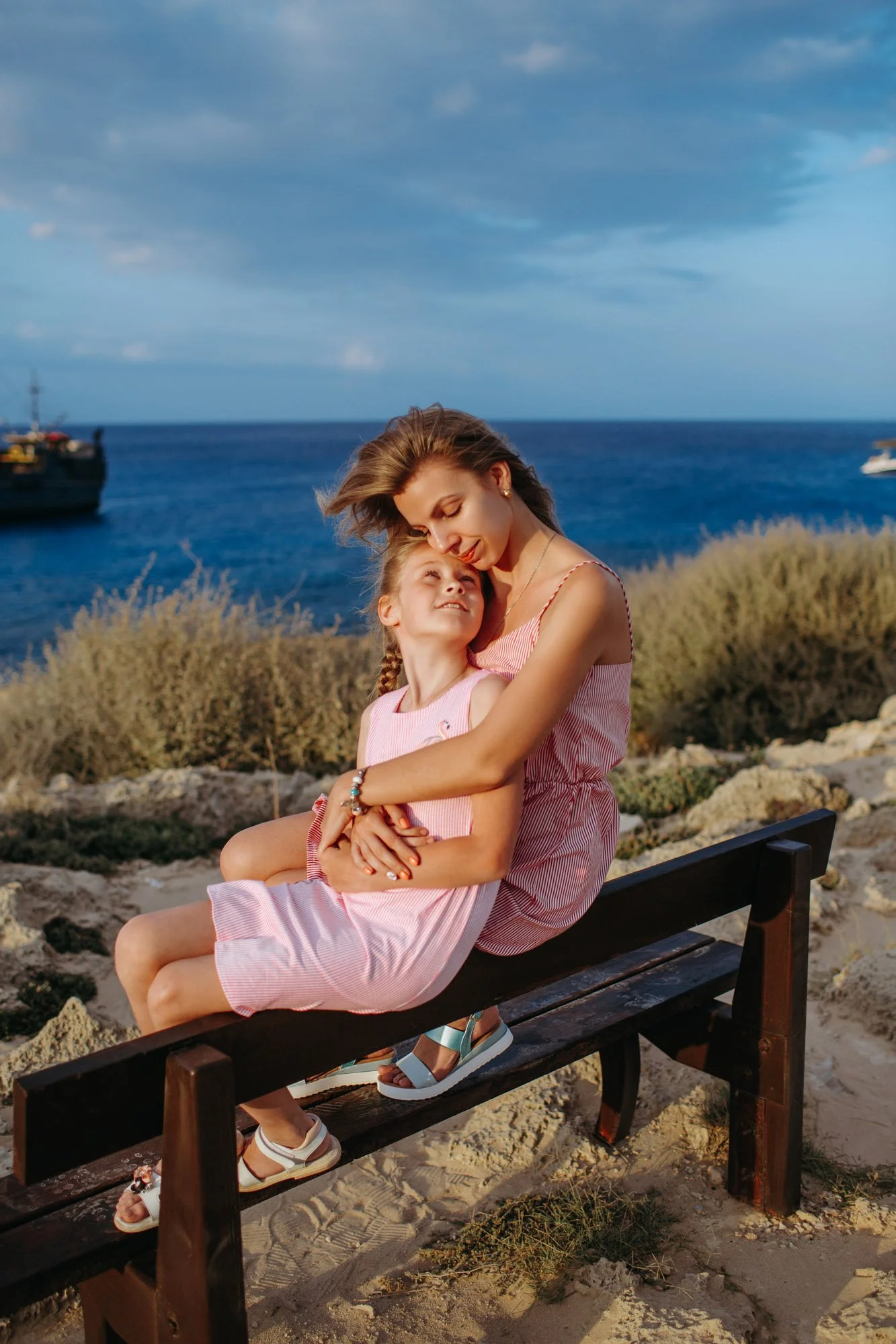 A woman and a young girl sitting on a park bench near the ocean, hugging, with boats in the water and a cloudy sky overhead.