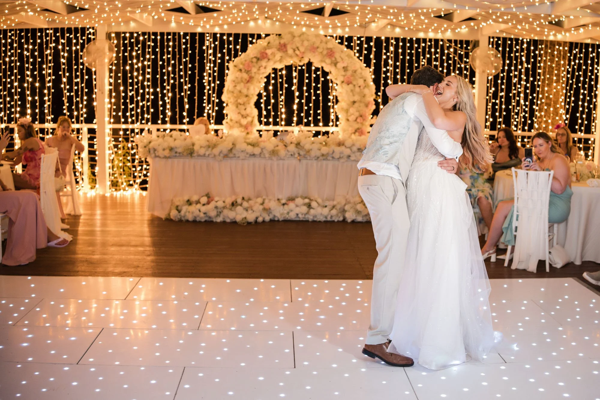 A couple dances together at a wedding reception under string lights and a flower arch, surrounded by seated guests.
