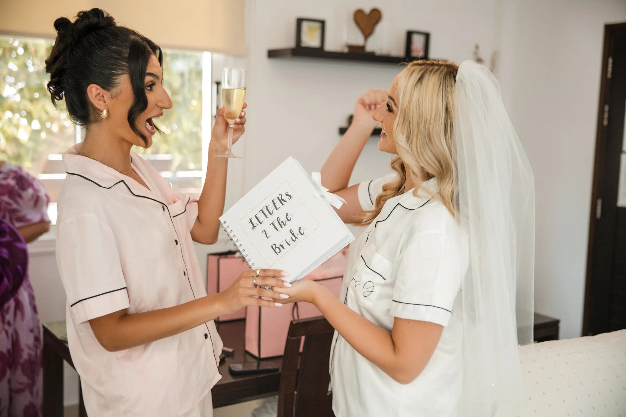 Two women in pajamas sharing a laugh, one holding a champagne glass and the other holding a wedding letter book, in a decorated room, possibly during a bridal celebration.