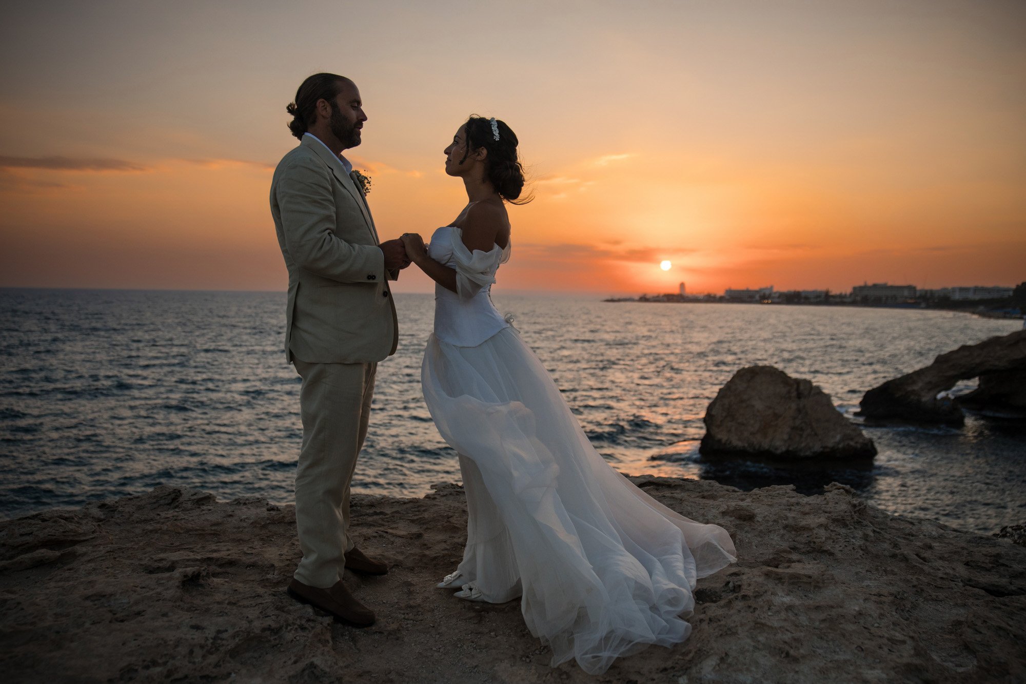 A couple dressed in wedding attire holding hands on a rocky coastline at sunset, with the ocean in the background.