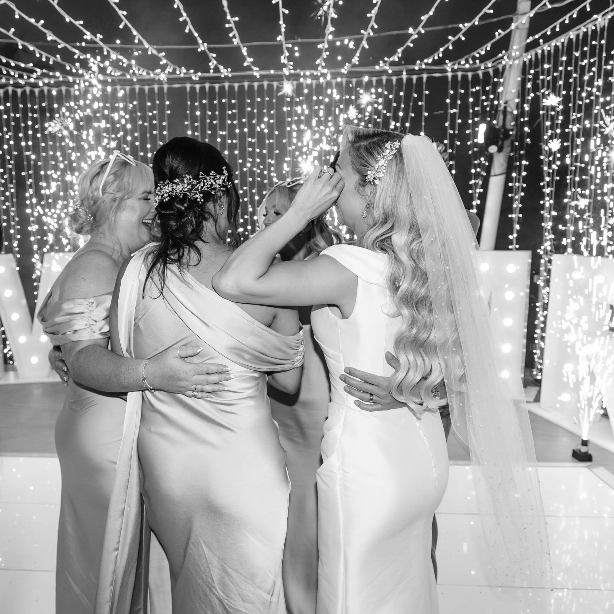 A group of women, including a bride with a veil, sharing a joyful moment at a wedding reception with sparklers and decorative lights in the background.