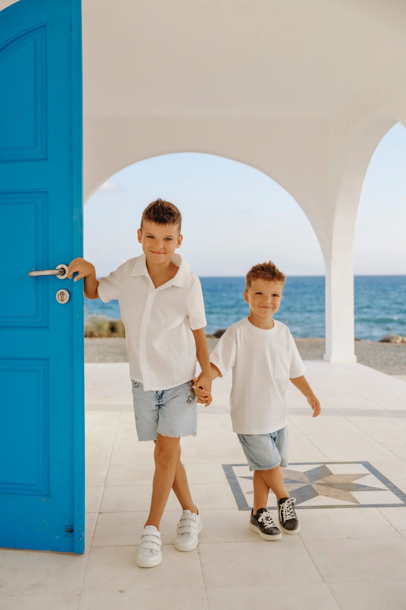 Two young boys holding hands, standing in the doorway of a building with a view of the ocean in the background.