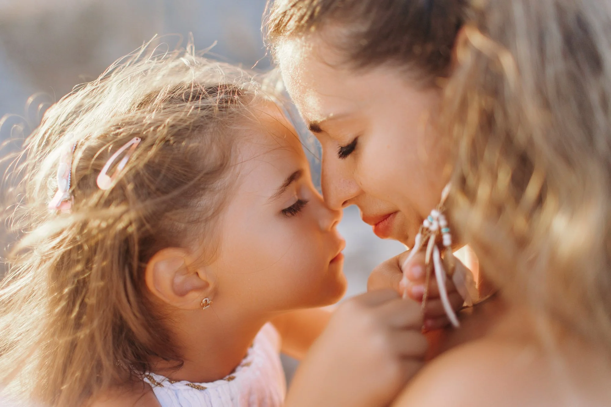 A woman and a young girl with curly hair share a tender moment with their foreheads touching and eyes closed, outdoors in natural light.