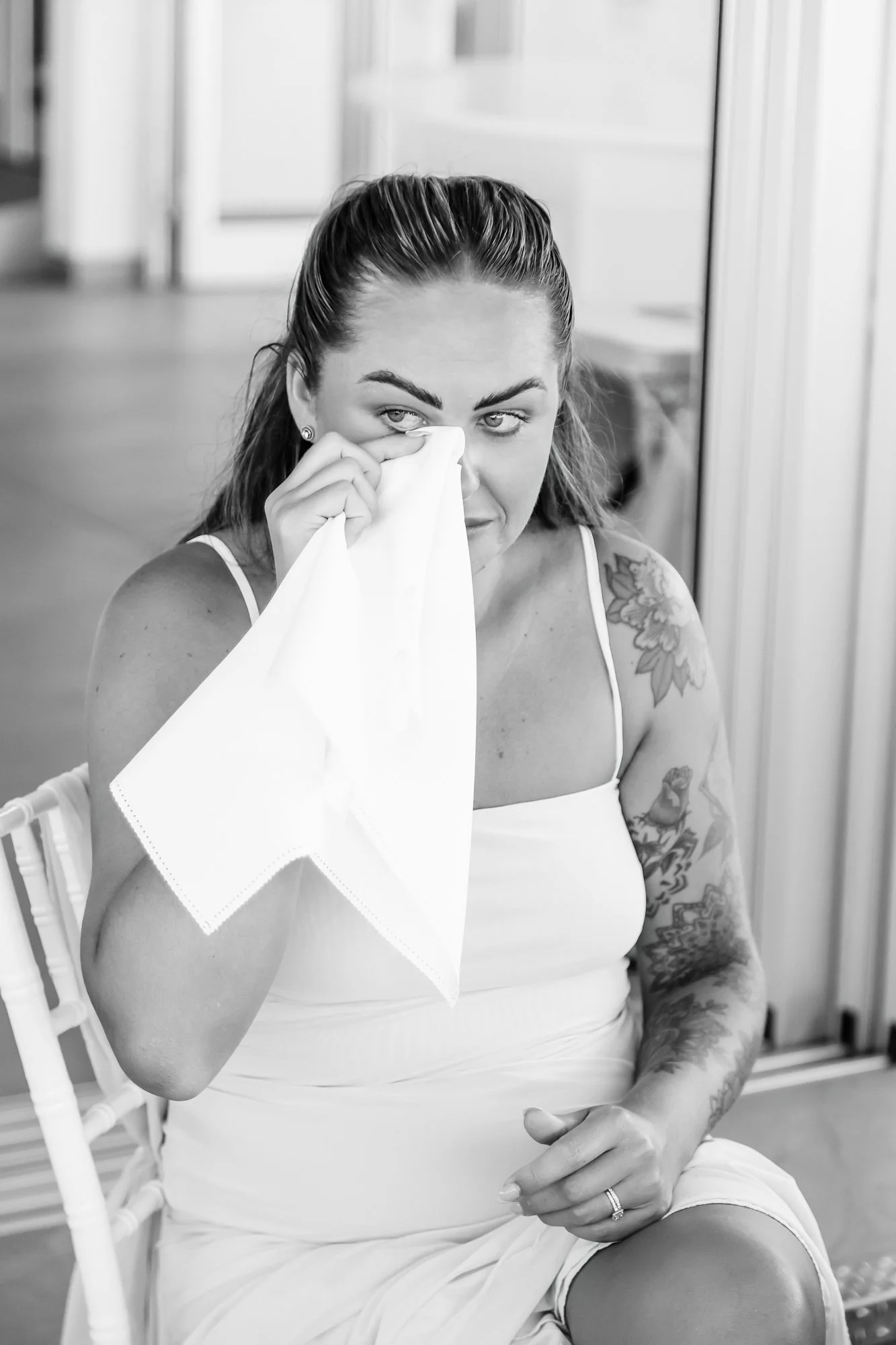 A woman with tattoos sitting on a chair, holding a tissue to her eye and wiping it, wearing a sleeveless dress.