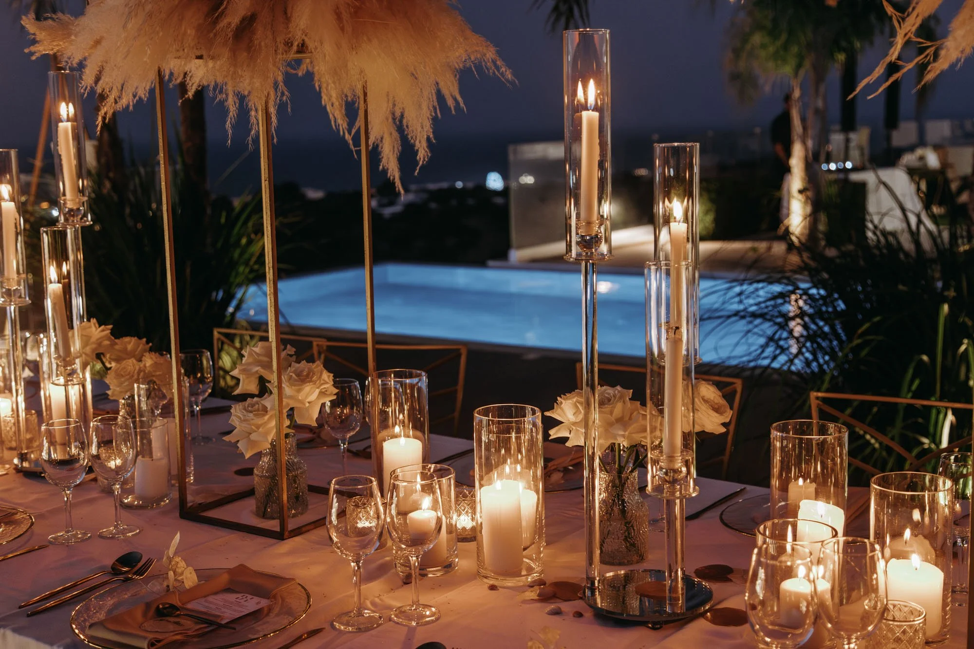 Nighttime outdoor dining table decorated with candles, flowers, and tall glass candle holders, overlooking a pool and ocean view.