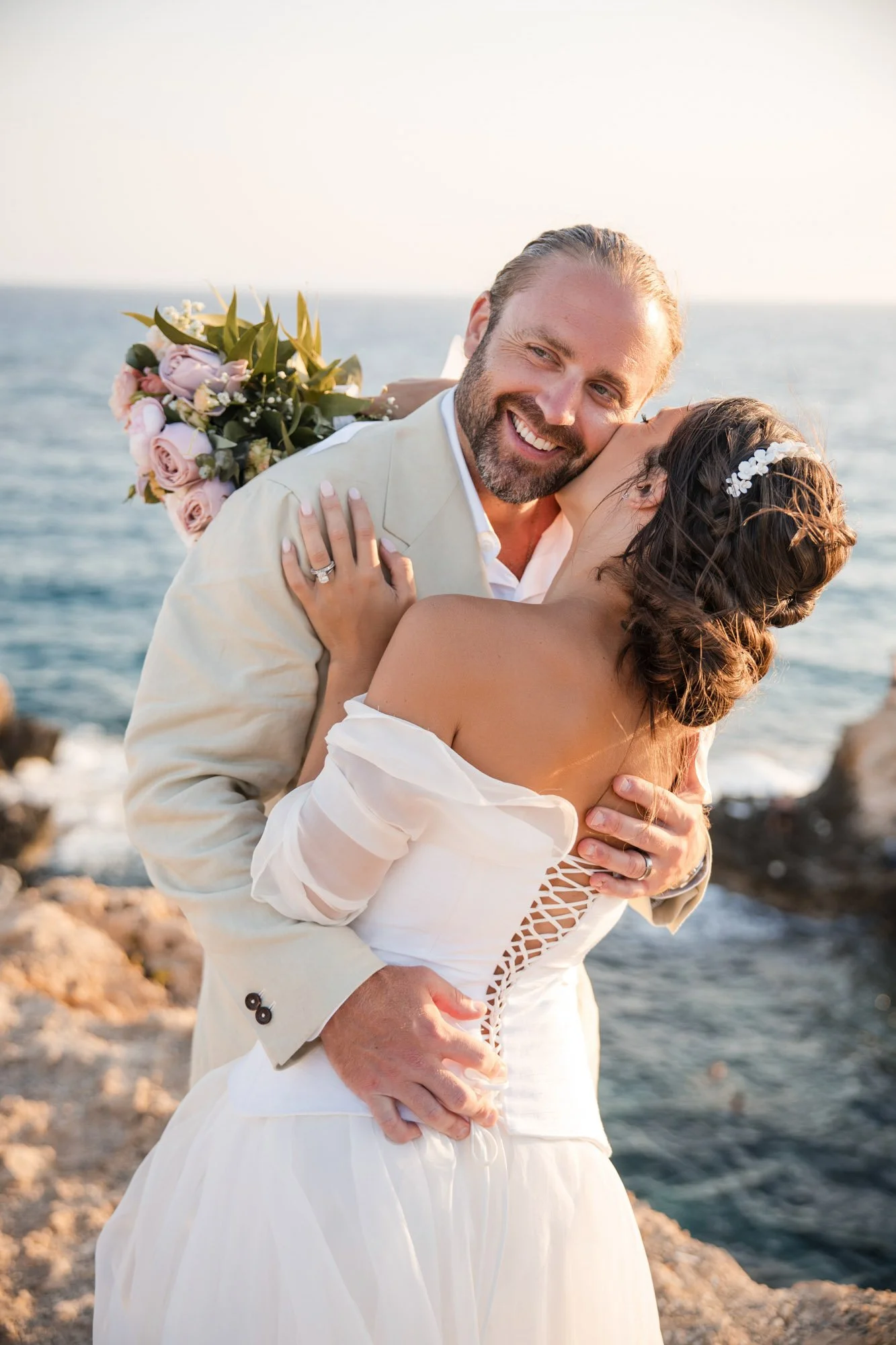 A newlywed couple embracing on a rocky shore by the ocean. The groom is smiling, holding a bouquet of pink and white flowers behind his back, while the bride is kissing his cheek. The bride wears a white off-the-shoulder wedding dress with lace-up ba