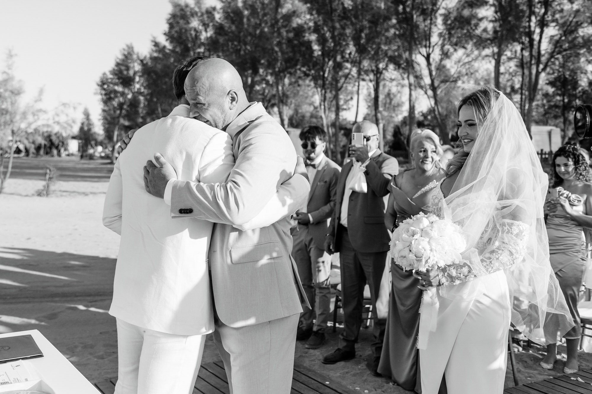 People hugging and smiling during a wedding ceremony outdoors with a bride holding a bouquet of flowers and guests capturing the moment.