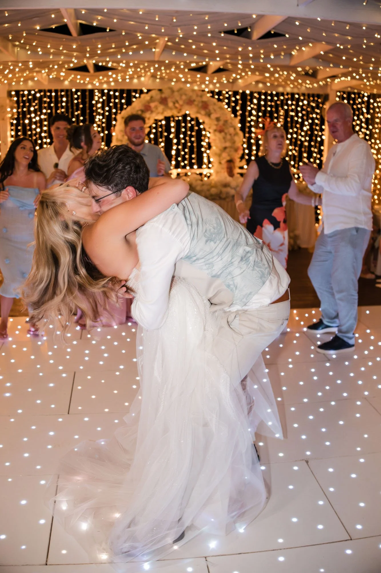 A bride and groom dance closely at their wedding reception, surrounded by guests, with string lights and floral decorations in the background.