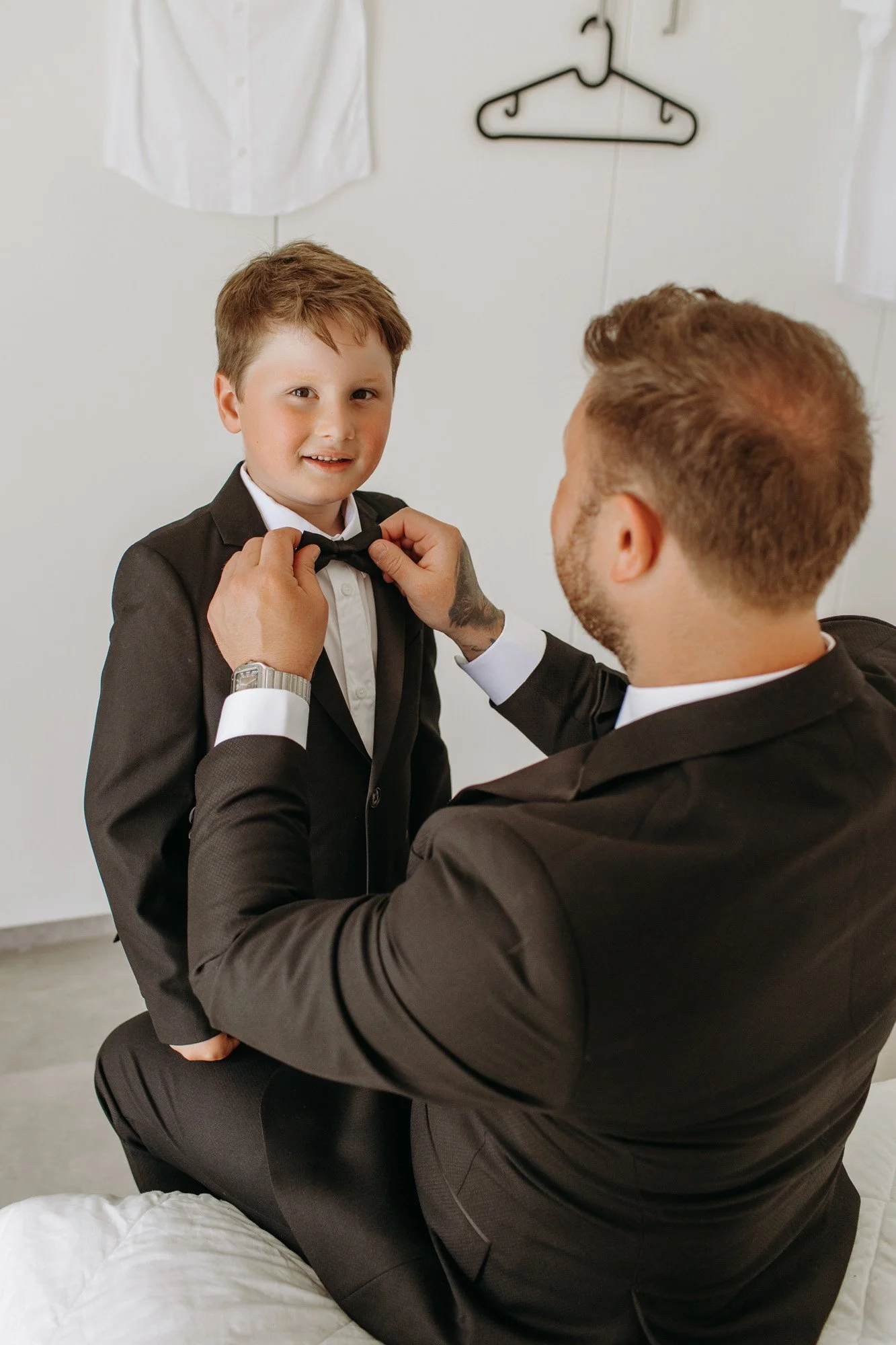 A man helping a young boy adjust his black bow tie in a bedroom with white walls and clothes hanging on a hook.