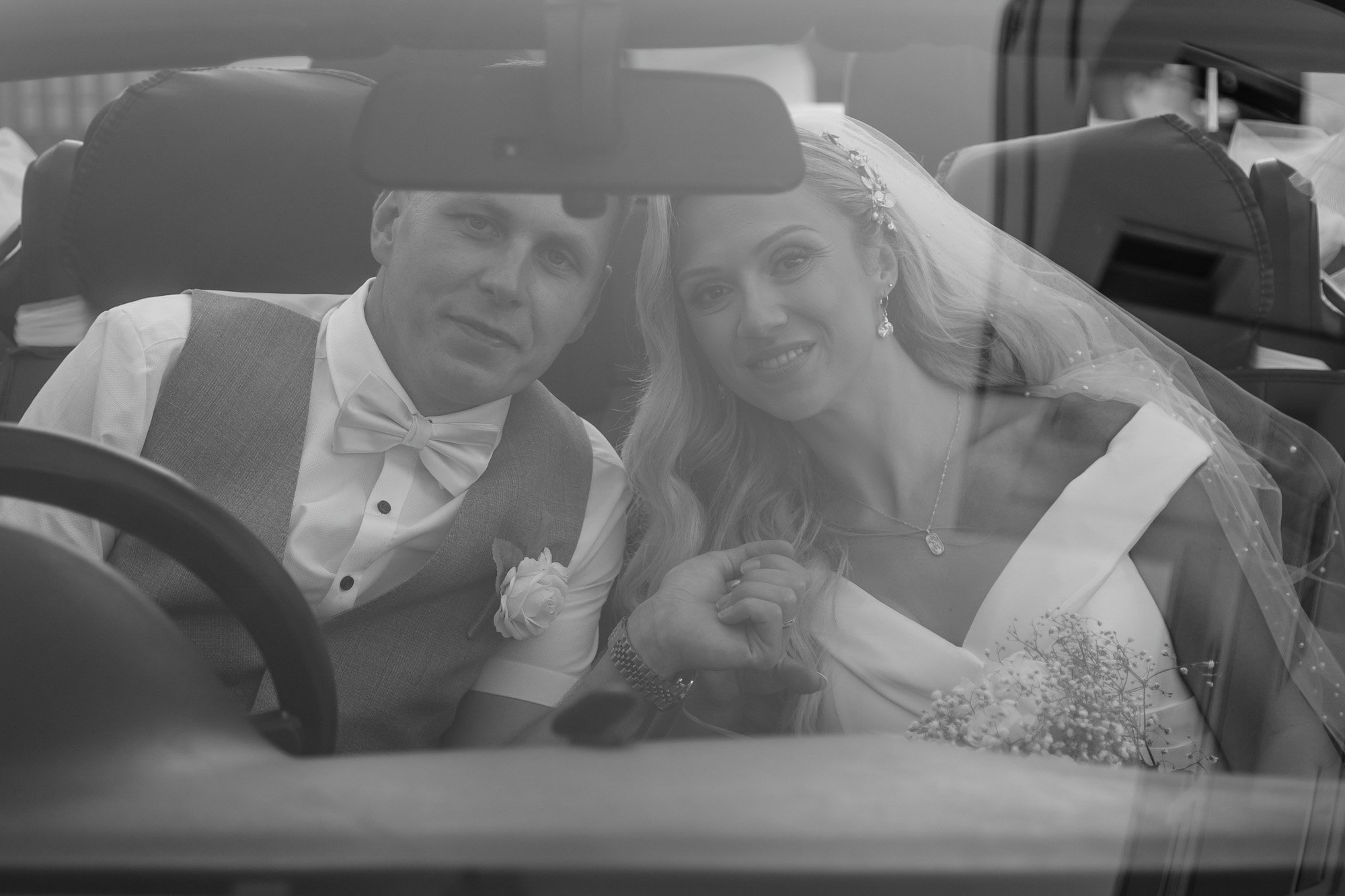 Black and white photo of a bride and groom sitting inside a car, looking through the windshield, with the bride wearing a veil and holding a bouquet.