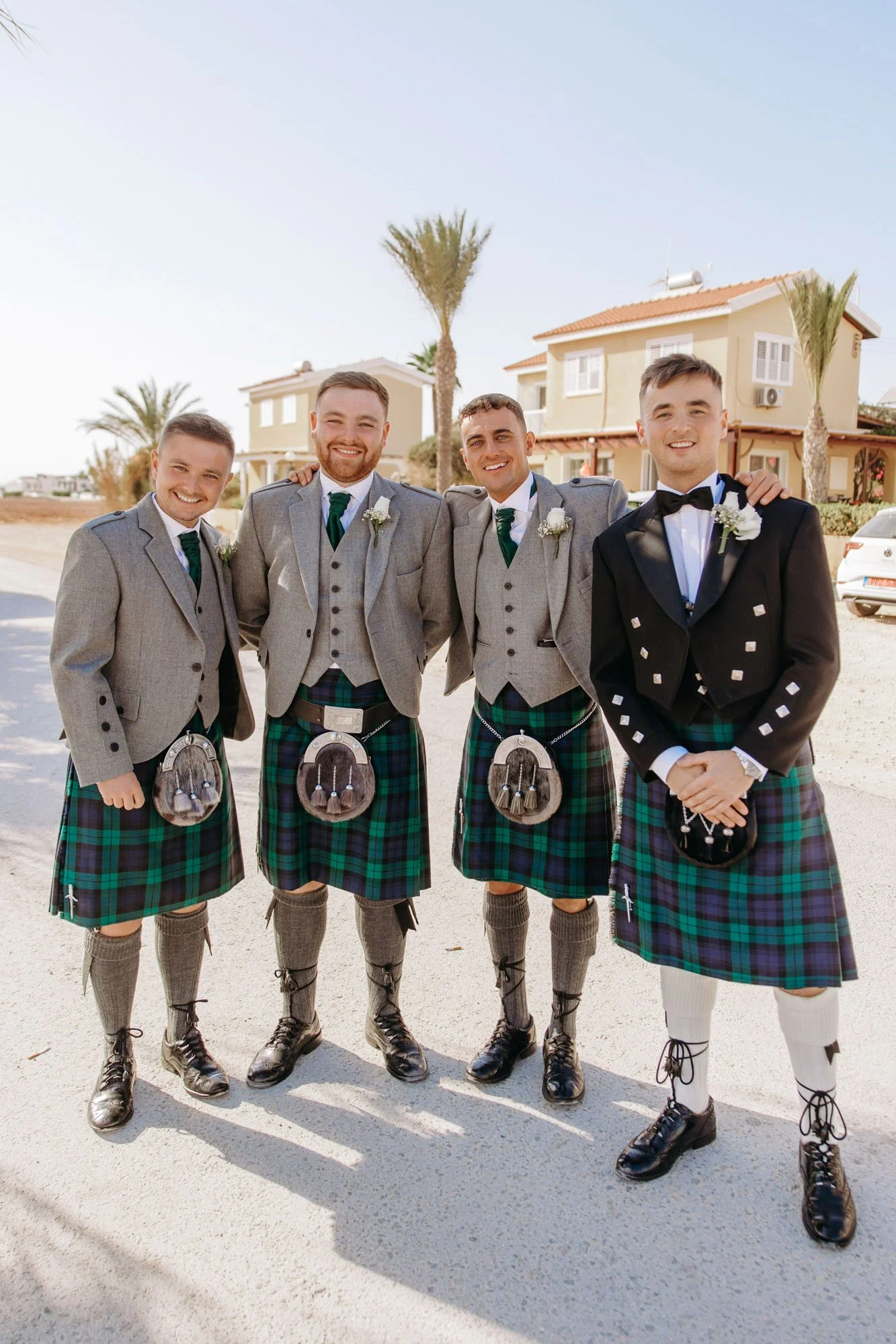 Four men in traditional Scottish attire, standing outdoors on a sunny day, smiling at the camera. They are wearing kilts with tartan patterns, jackets, and accessories, with one in a formal black jacket and bow tie. Residential houses and palm trees 