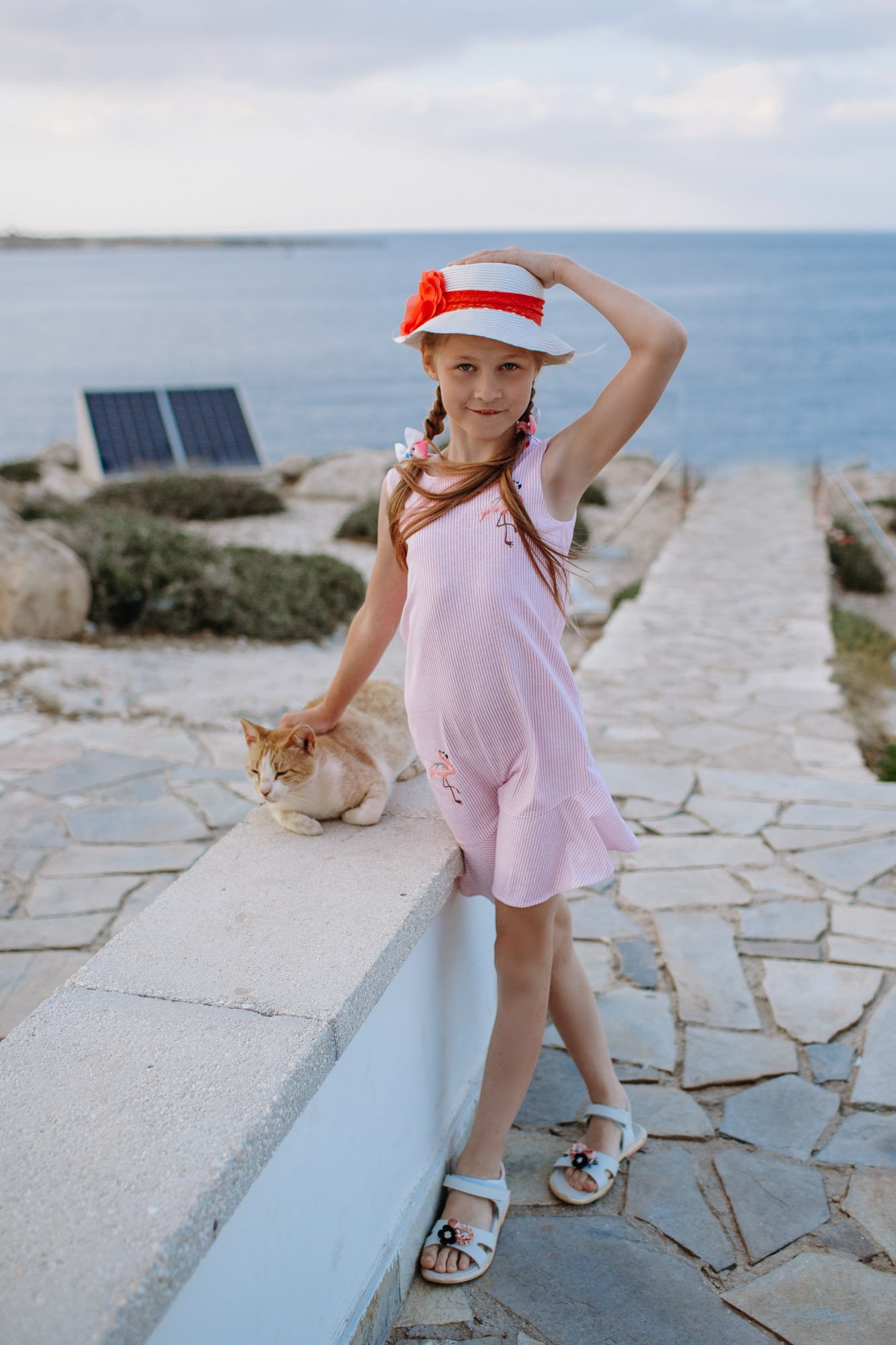 A young girl wearing a pink dress, a white sunhat with a red band and flower decoration, and sandals, standing outdoors near a stone wall and a cat, with a view of the ocean and cloudy sky in the background.
