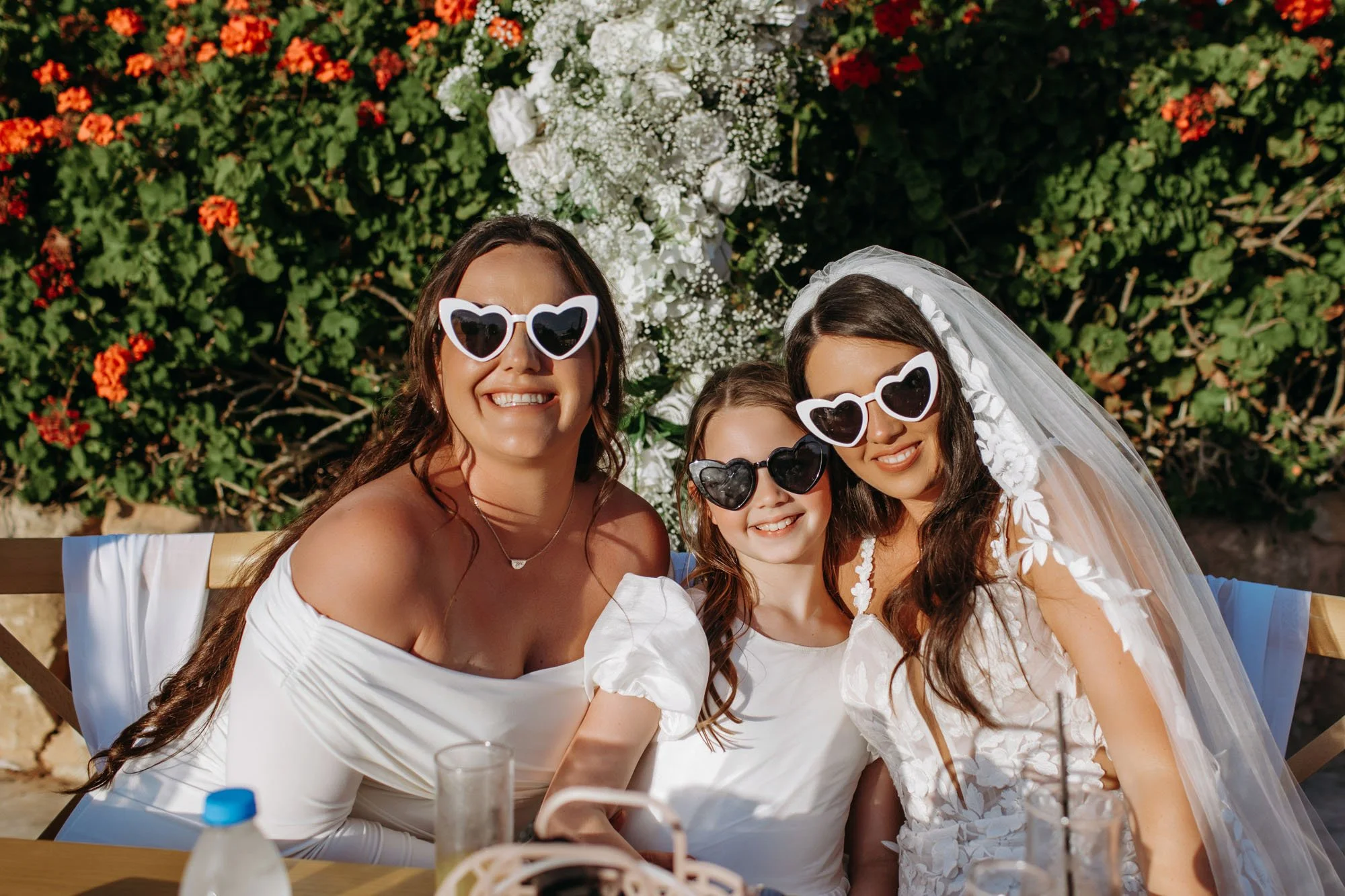 Three women, including a bride in a wedding dress with a veil, sitting outdoors with two girls, all wearing heart-shaped sunglasses, smiling for a photo during a celebration or wedding reception.