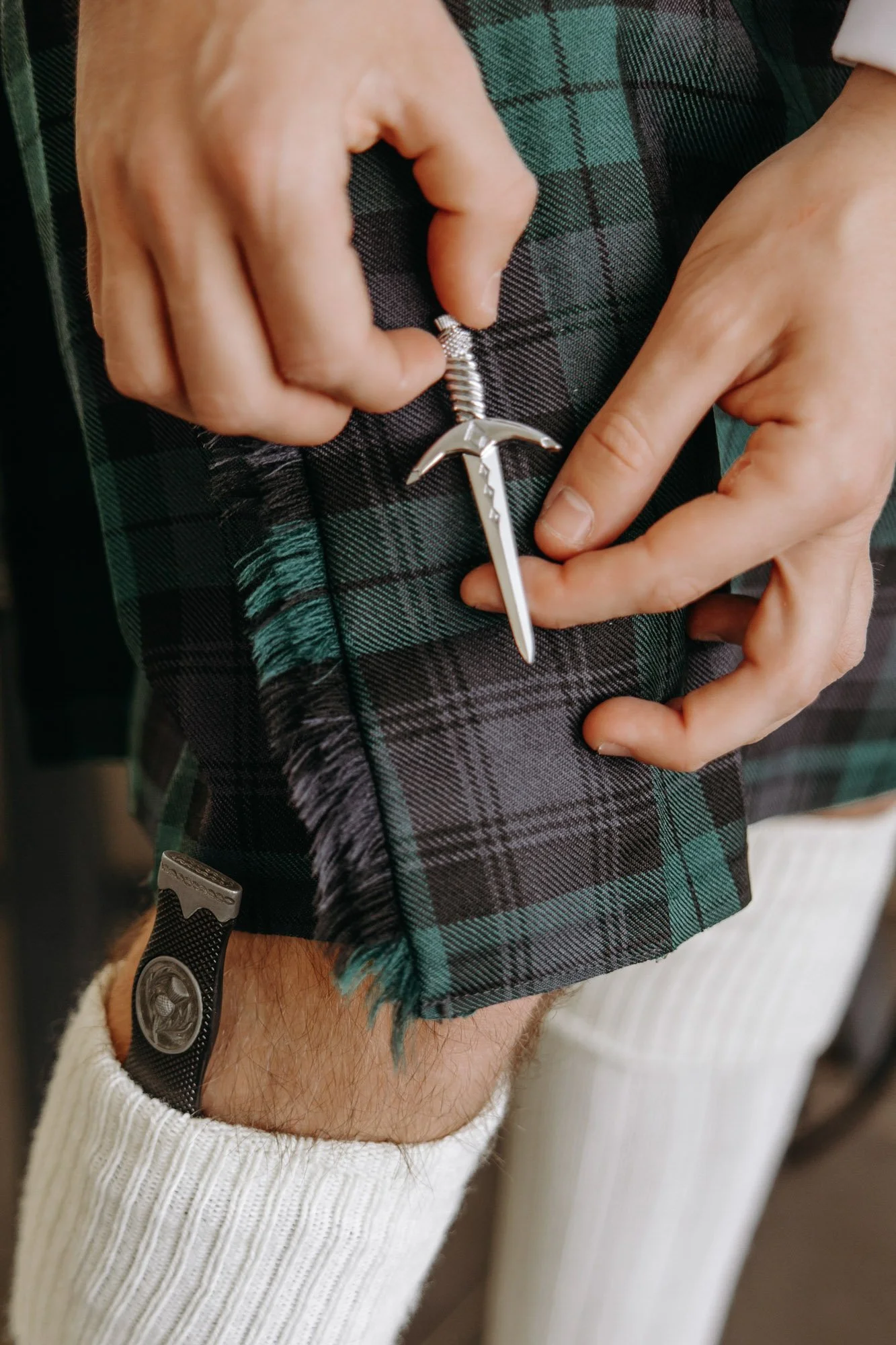 Person adjusting a silver anchor pendant necklace with their right hand, wearing tartan pants and a white sleeve. The person has a watch on their left wrist, which is visible under rolled-up pants.
