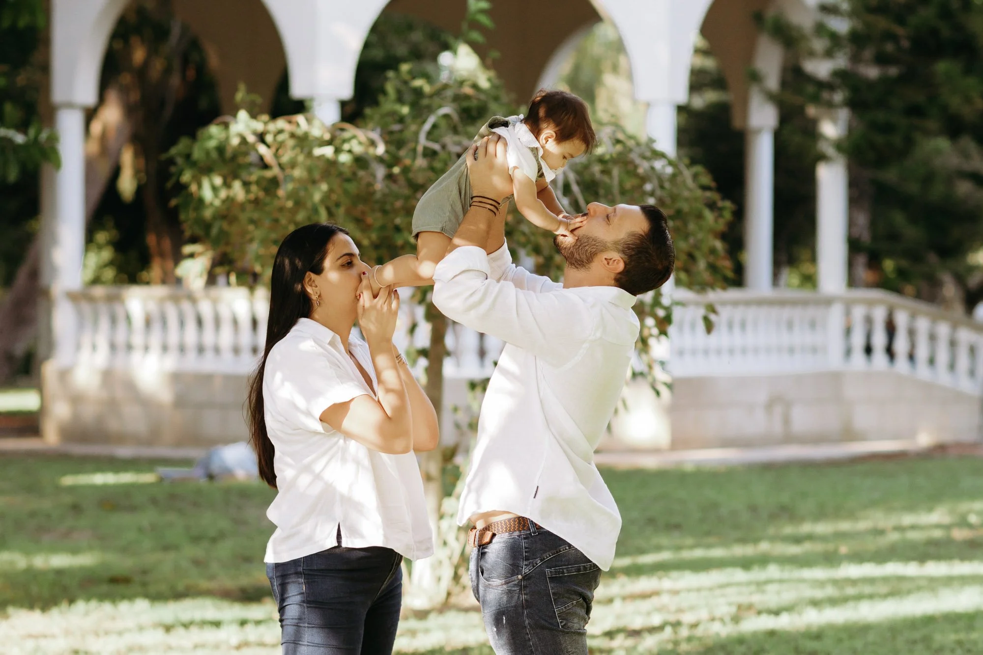 A family with a man, woman, and young child enjoying time together outdoors in a park or garden setting, with the man lifting the child and the woman holding the child's foot to her face.