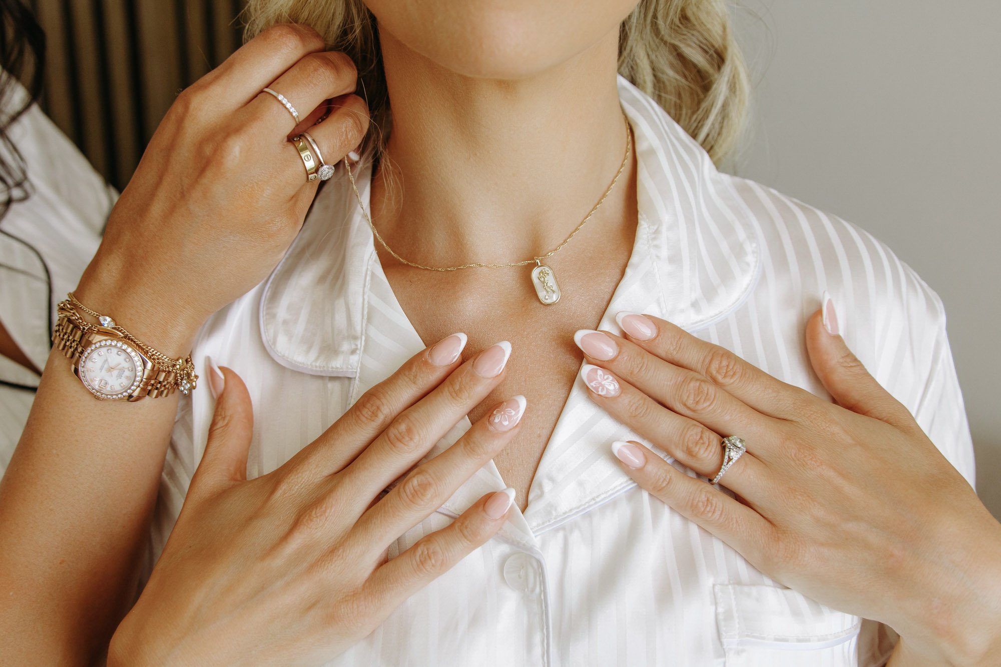 A woman wearing jewelry, including a gold watch, rings, and necklaces, with her hands gently touching her chest and face.