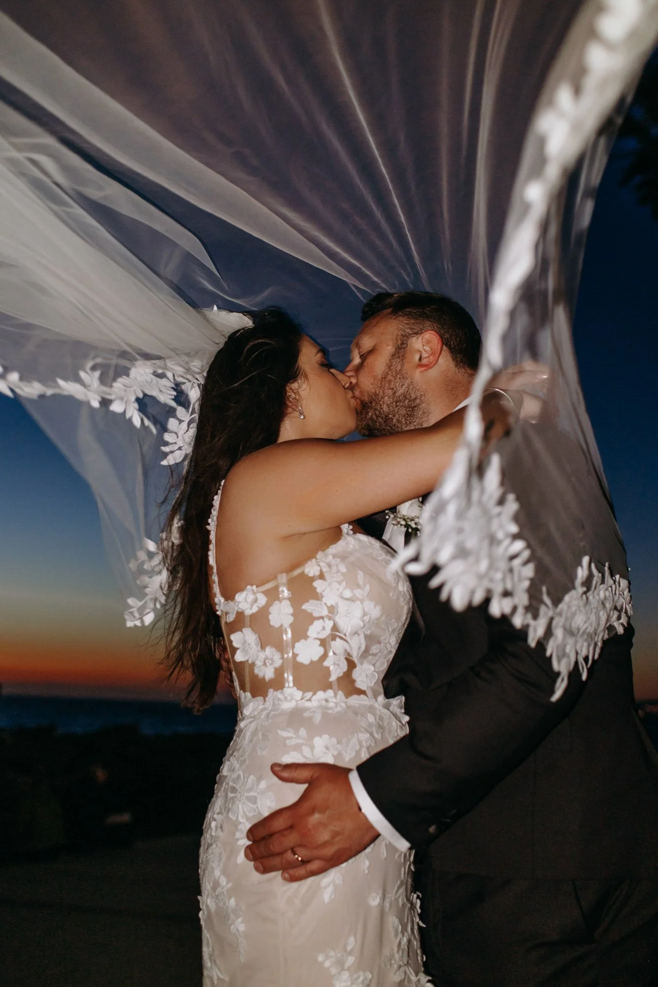 A bride and groom kissing under a veil at sunset by the ocean.