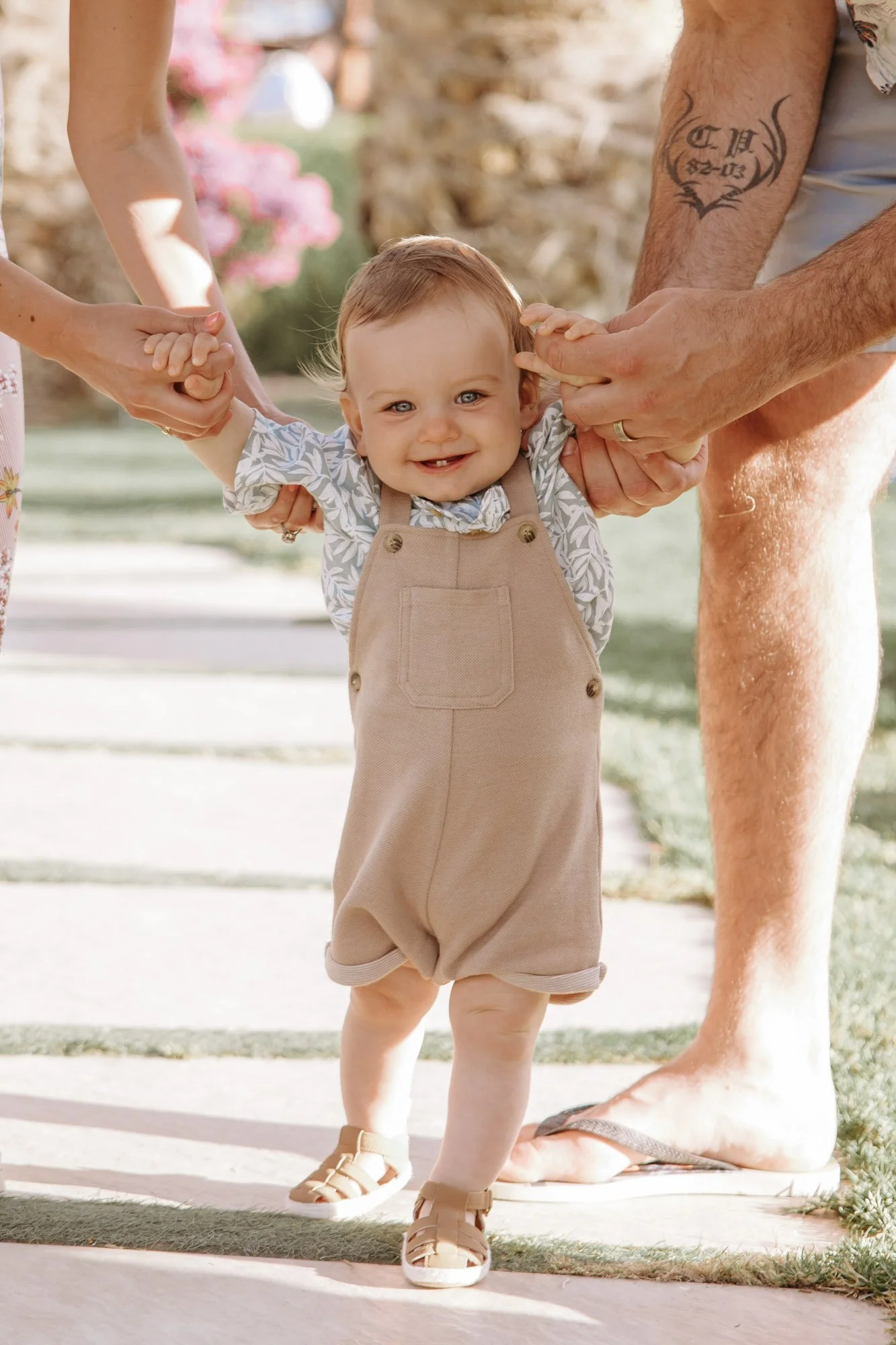 A toddler with light brown hair, blue eyes, and a big smile is walking outdoors while holding hands with two adults. One adult's arm with a tattoo is visible on the right, and the other adult is on the left. The child is wearing tan overalls, a patte