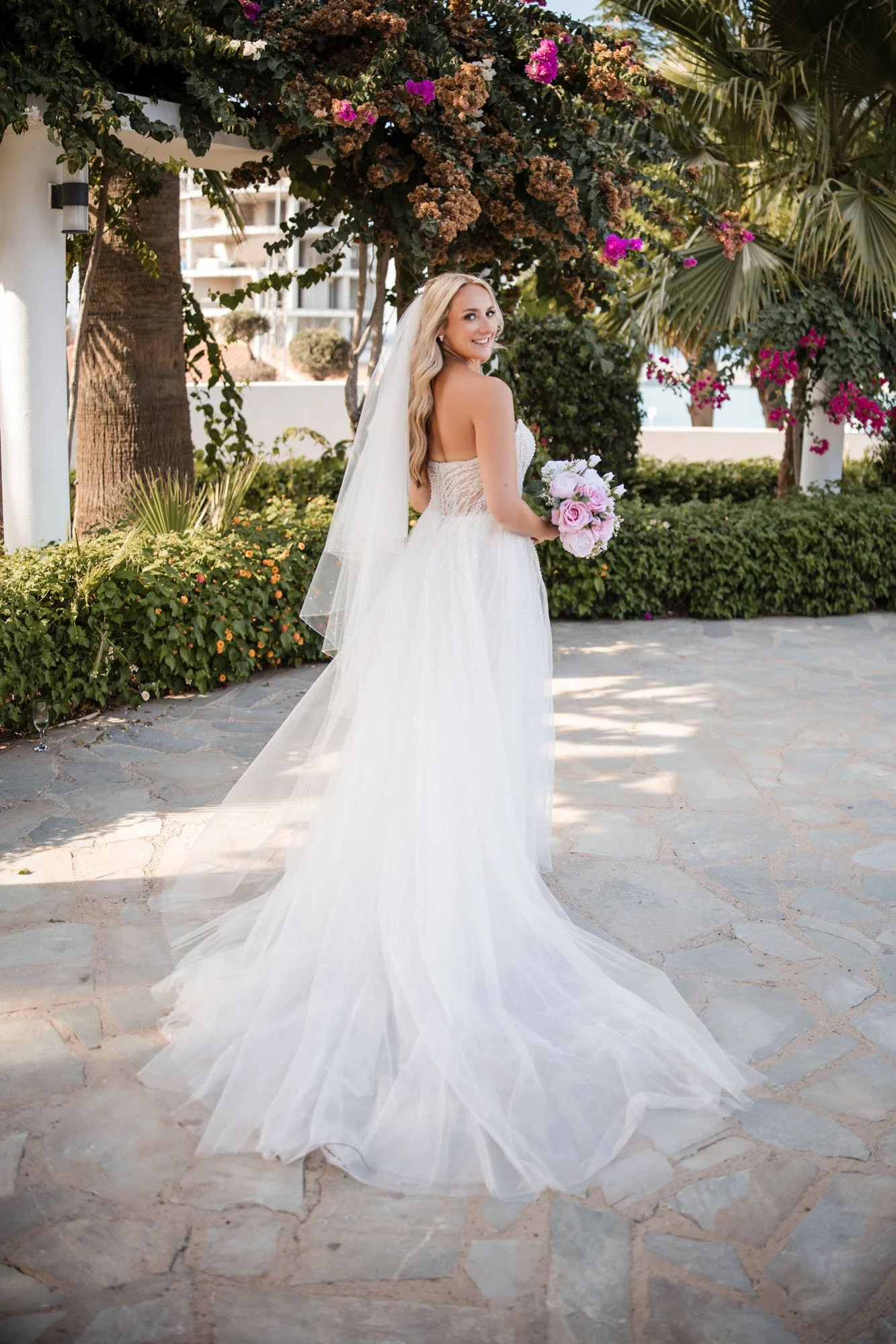 A bride in a strapless white wedding gown holding a bouquet of pink and white flowers, outside on a stone-paved area, under a flowering tree with pink blossoms and lush greenery.