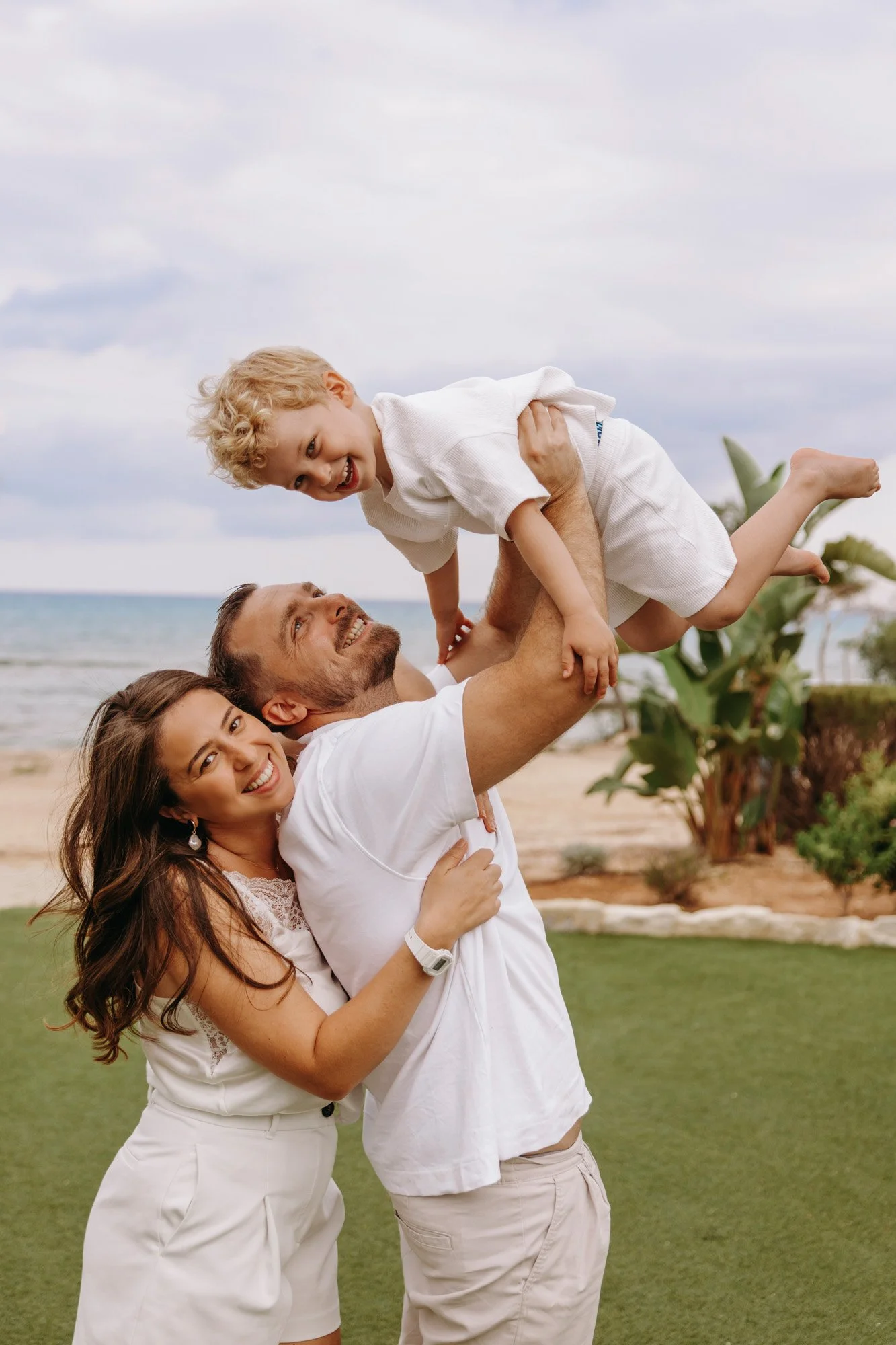 A family of four enjoying a day at the beach, with a woman and man lifting a young boy into the air while another woman looks on, all smiling.