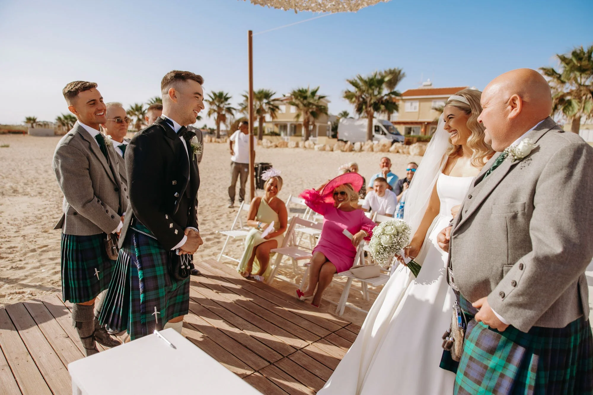 A wedding ceremony on the beach with a bride and groom facing each other, surrounded by friends and family. The bride is holding a bouquet, and some guests are seated on white chairs, with palm trees and beach houses in the background.
