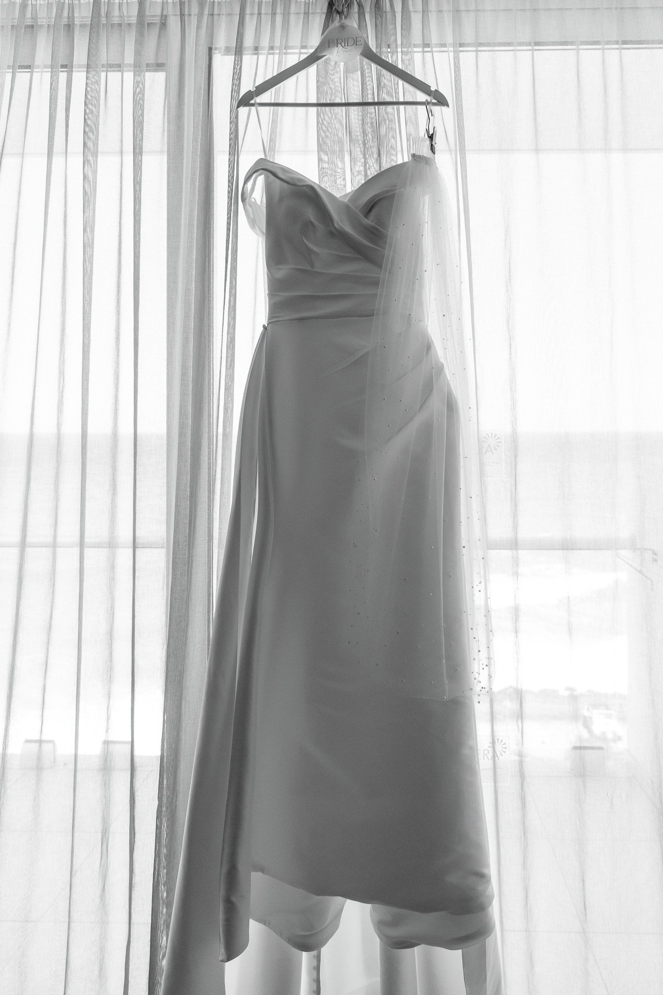 Wedding dress hanging on a hanger in front of a sheer curtain.