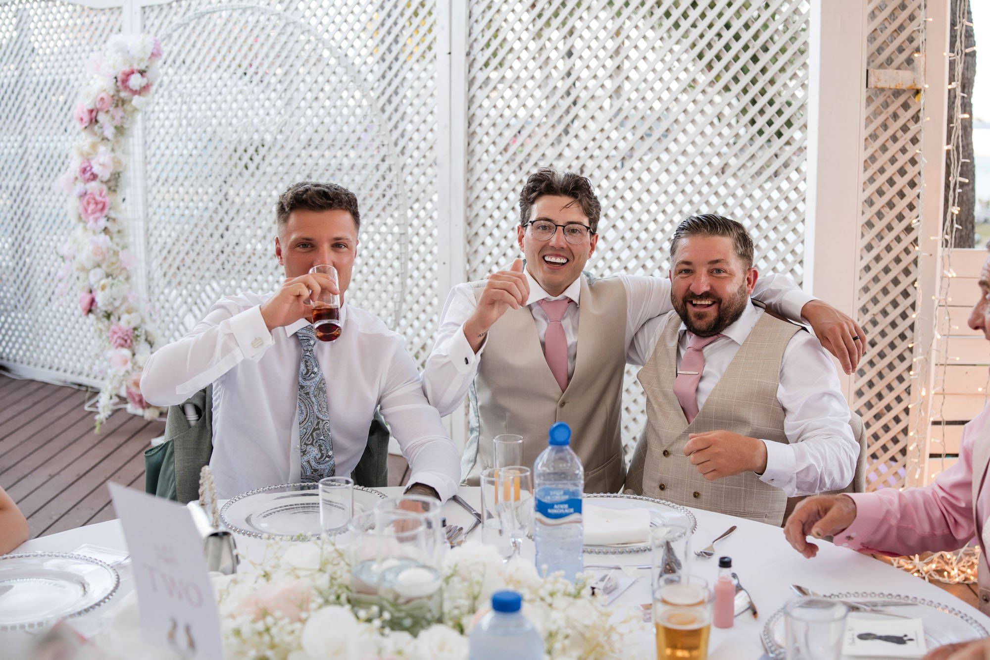 Three men seated at a decorated event table, smiling and enjoying drinks, with a braided floral arch in the background.
