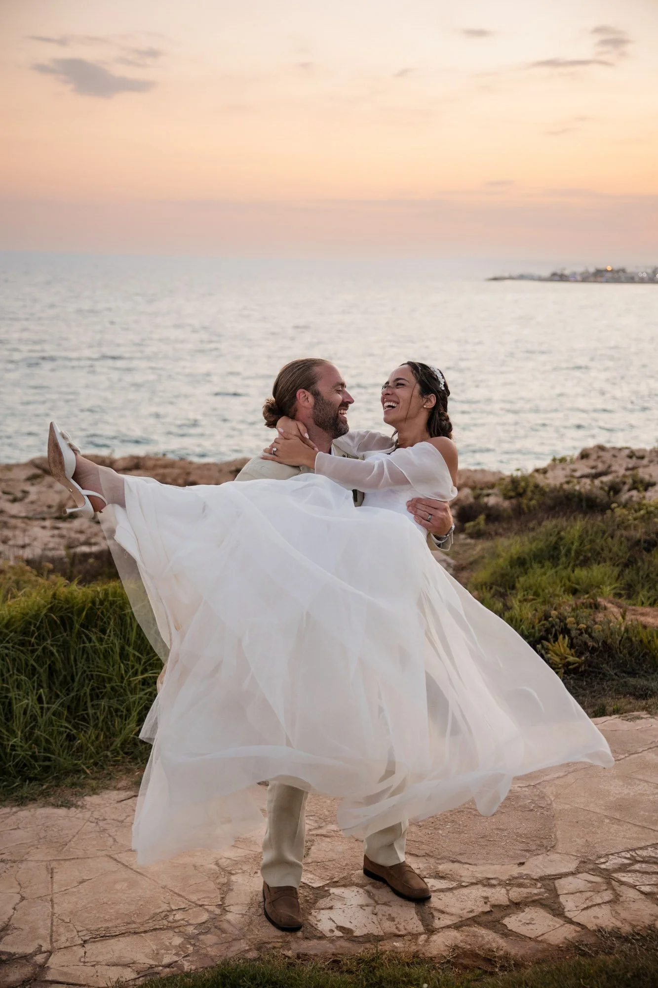 A man in a beige suit and a woman in a white wedding dress are at the coast during sunset. The man is holding the woman in his arms, and they are smiling and looking at each other.